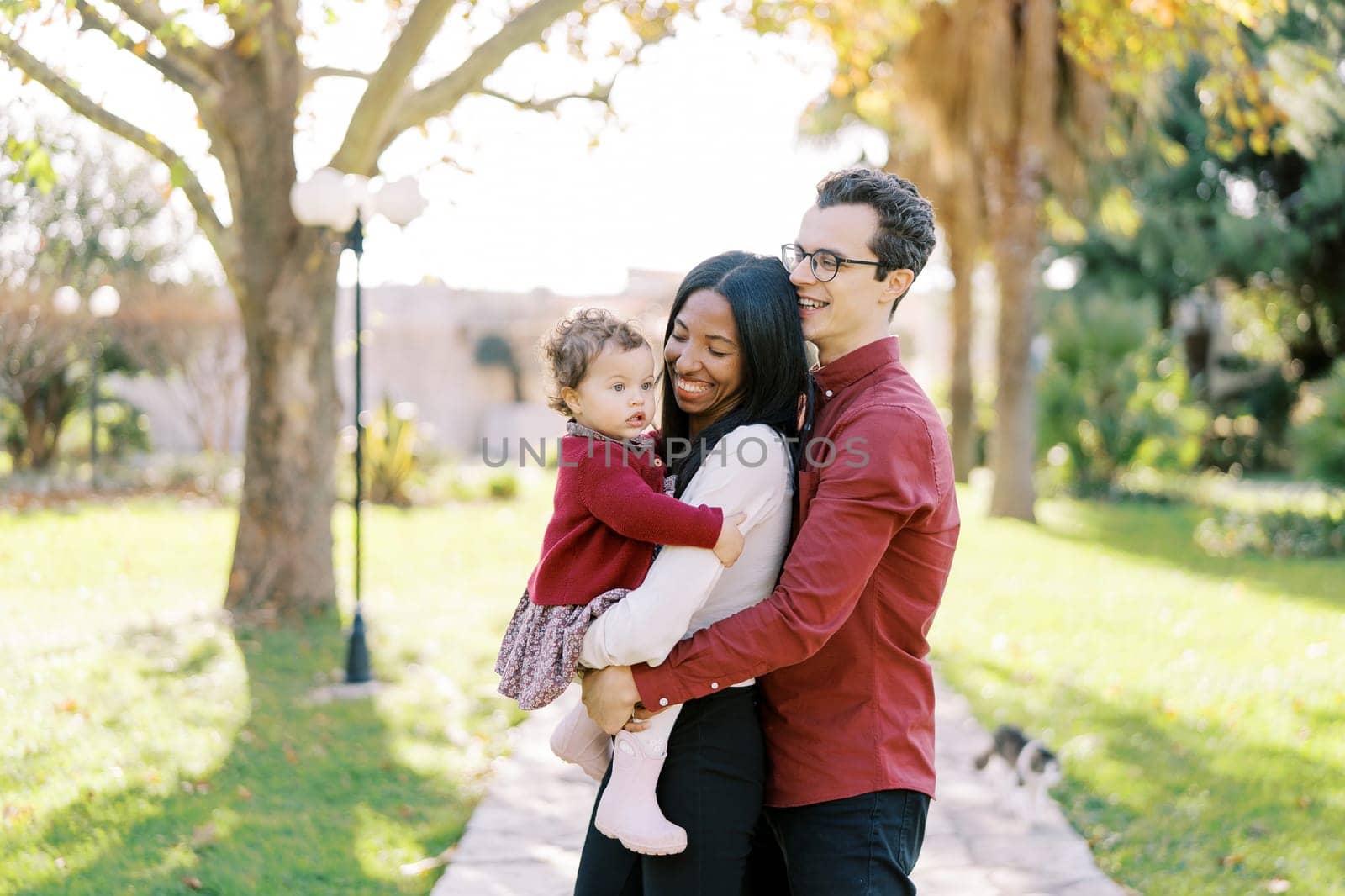 Smiling dad hugging mom from behind with a little girl in her arms in the sunny park. High quality photo