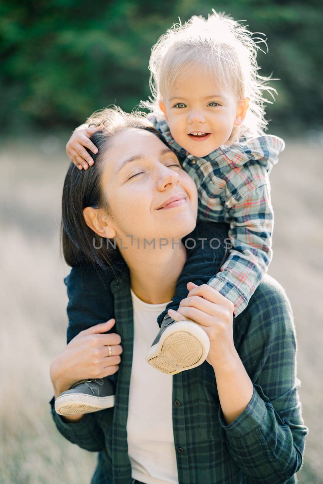 Happy little girl hugging her mom head while sitting on her shoulders. Portrait by Nadtochiy