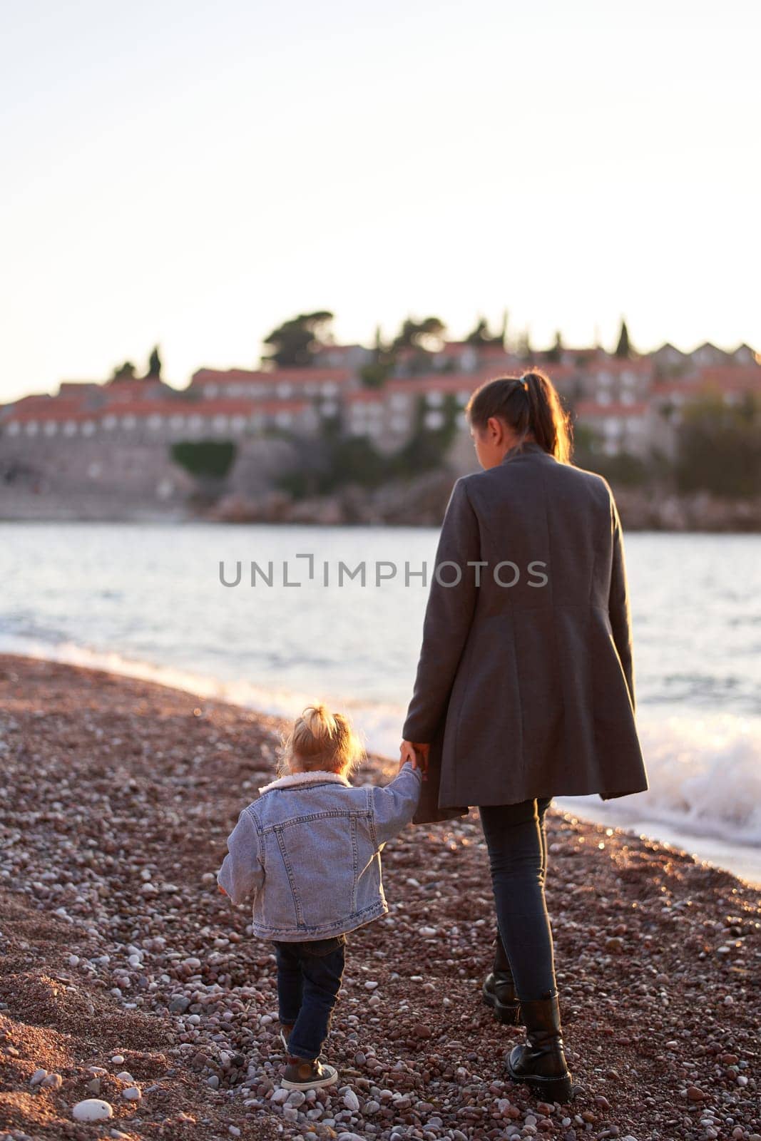 Mom and a little girl walk holding hands along the seashore to the island of Sveti Stefan. Back view by Nadtochiy