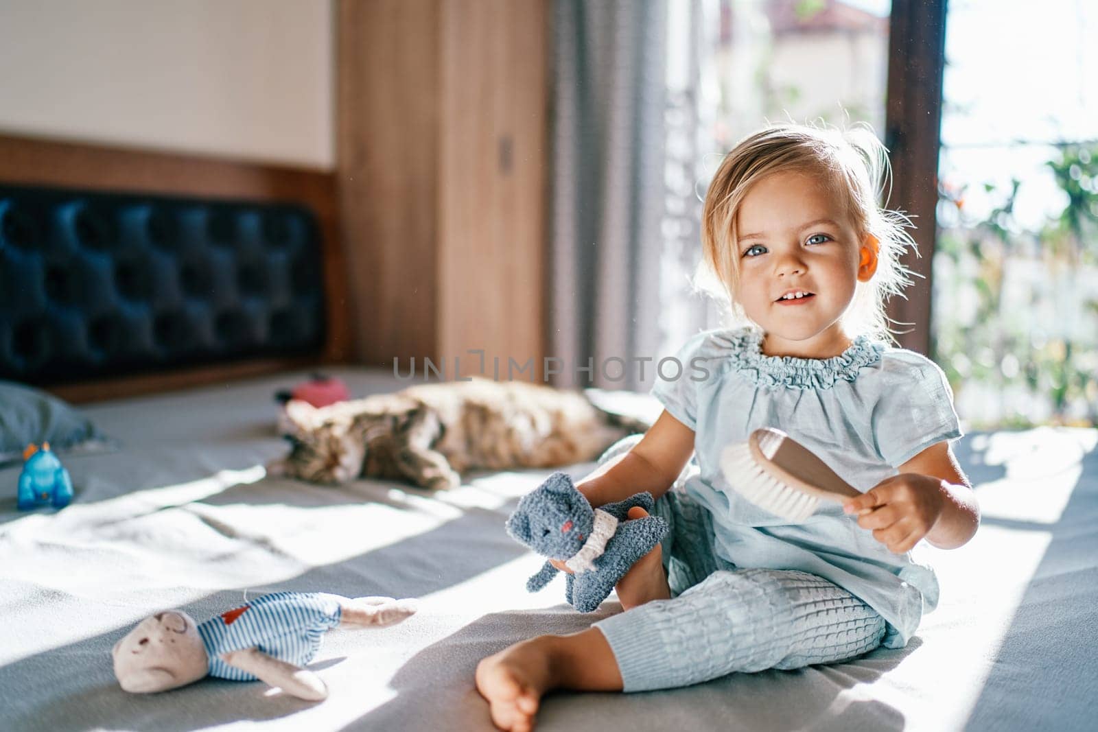 Little smiling girl sitting on the bed with a toy cat and a comb in her hands by Nadtochiy