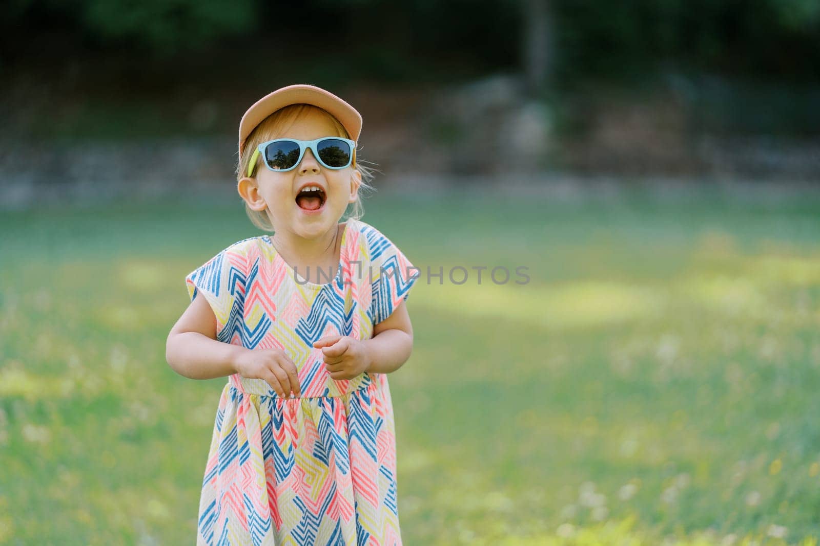 Little laughing girl stands in sunglasses on a sunny lawn by Nadtochiy