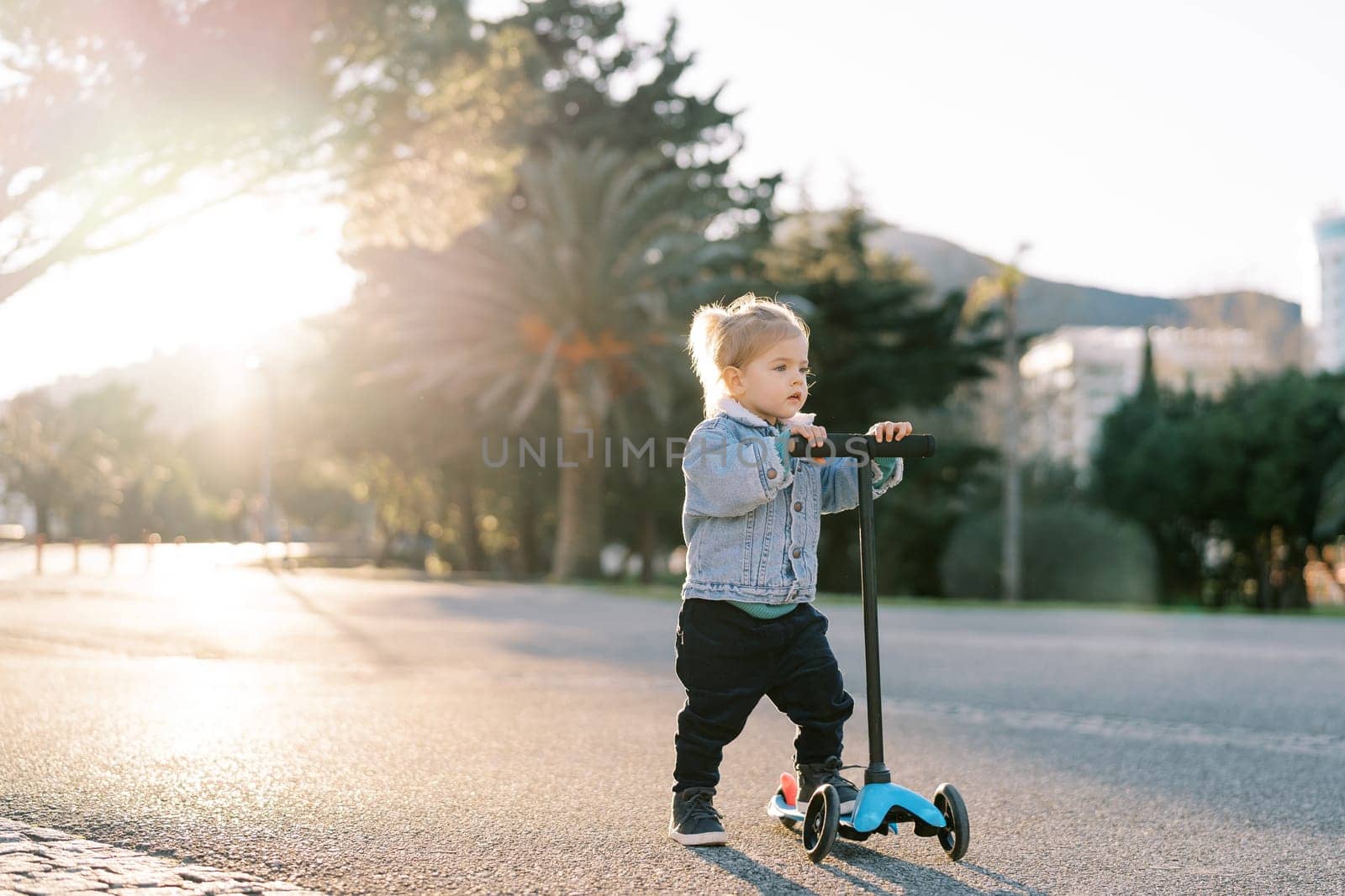 Little girl stands holding the steering wheel of a scooter on the road and looks ahead by Nadtochiy