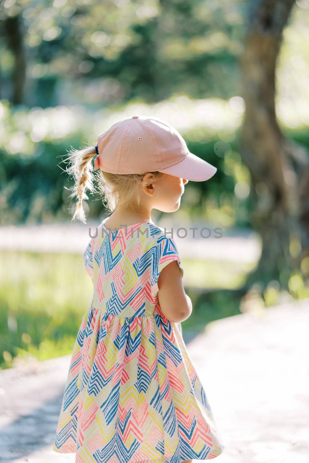 Little girl in a cap walks along a path in a sunny garden. Back view. High quality photo