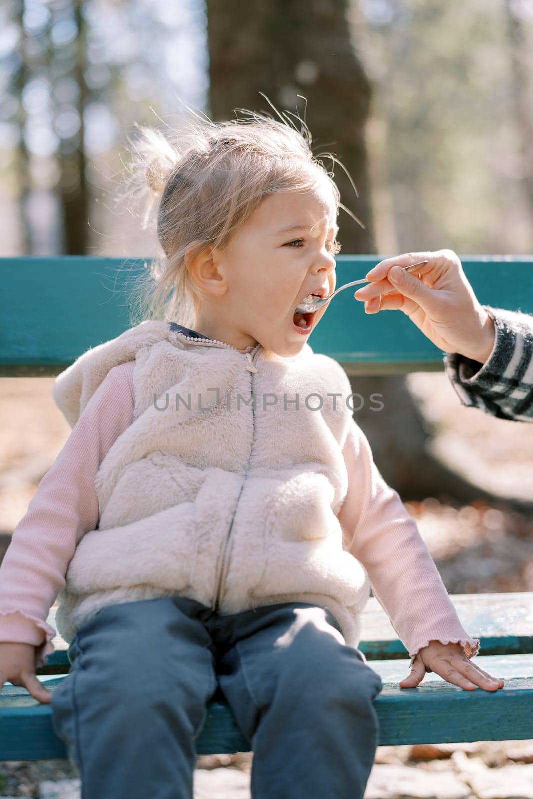 Little girl eats porridge from a spoon which her mother feeds her, sitting on a bench in the park. Cropped by Nadtochiy