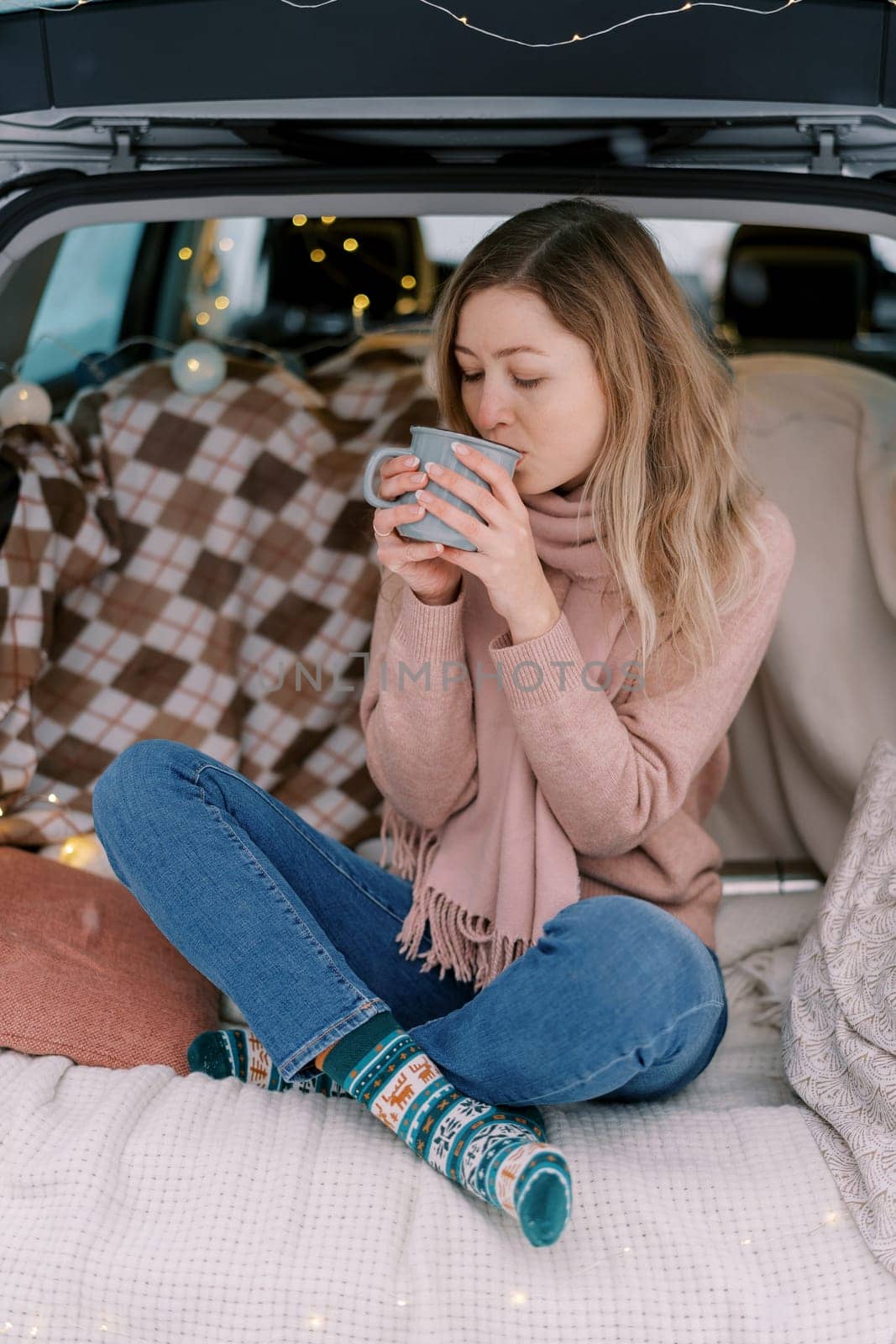 Girl sits cross-legged on a blanket in the trunk of a car and drinks coffee from a mug. High quality photo