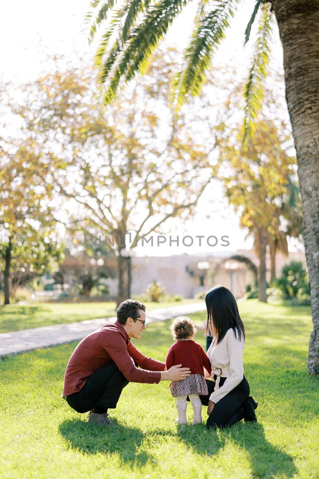Dad hugs a little girl squatting next to mom in the park. Back view. High quality photo