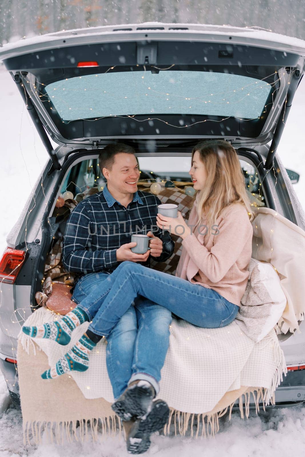 Girl put her feet on the legs of guy sitting with cups of coffee in the trunk of the car in the winter forest by Nadtochiy