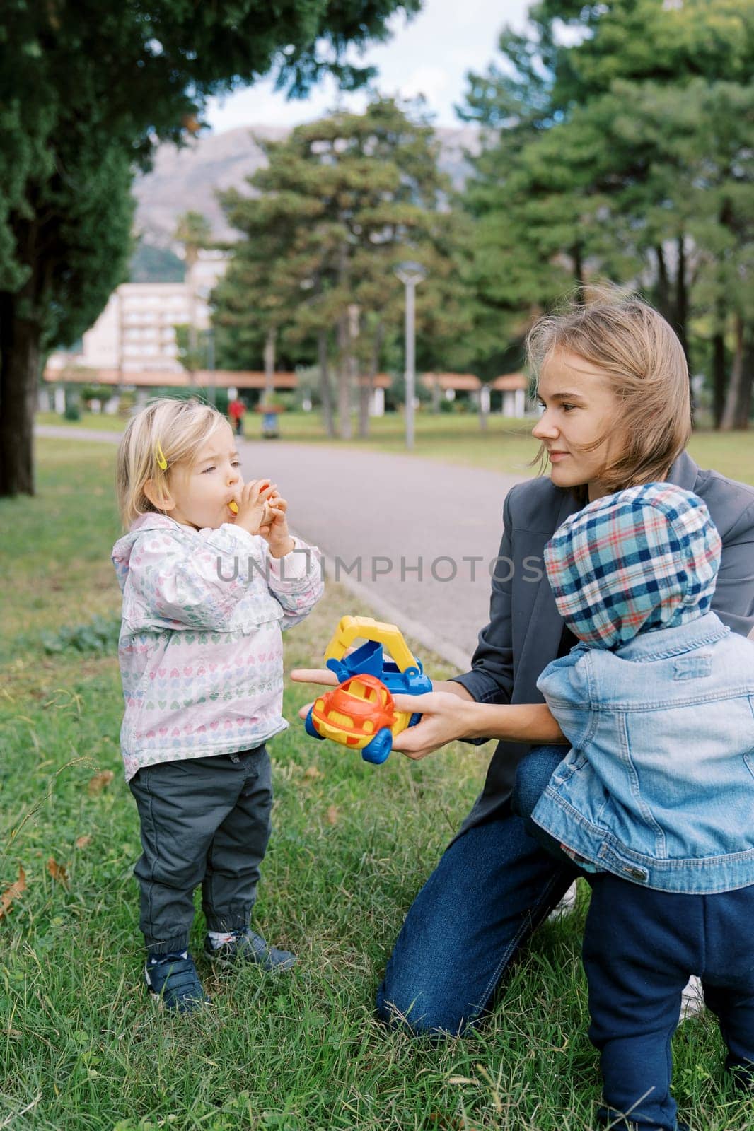 Small boy stands near his mother holding out toy cars to a little girl. High quality photo