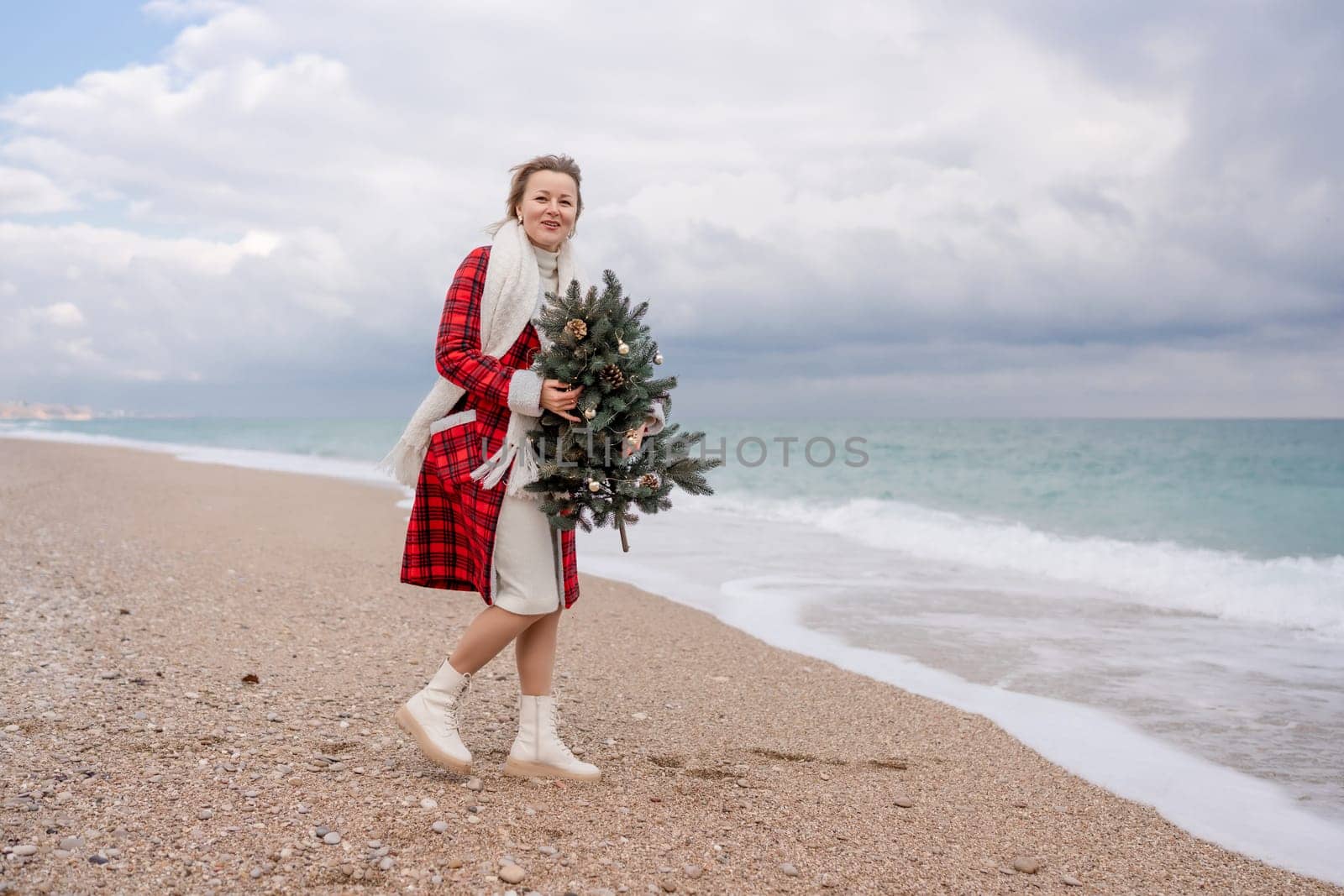 Blond woman holding Christmas tree by the sea. Christmas portrait of a happy woman walking along the beach and holding a Christmas tree in her hands. Dressed in a red coat, white dress