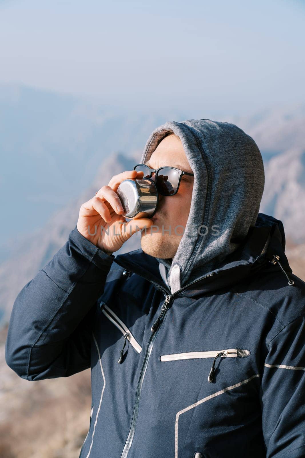 Man in a hooded jacket drinks coffee from a mug of a thermos in the mountains. High quality photo