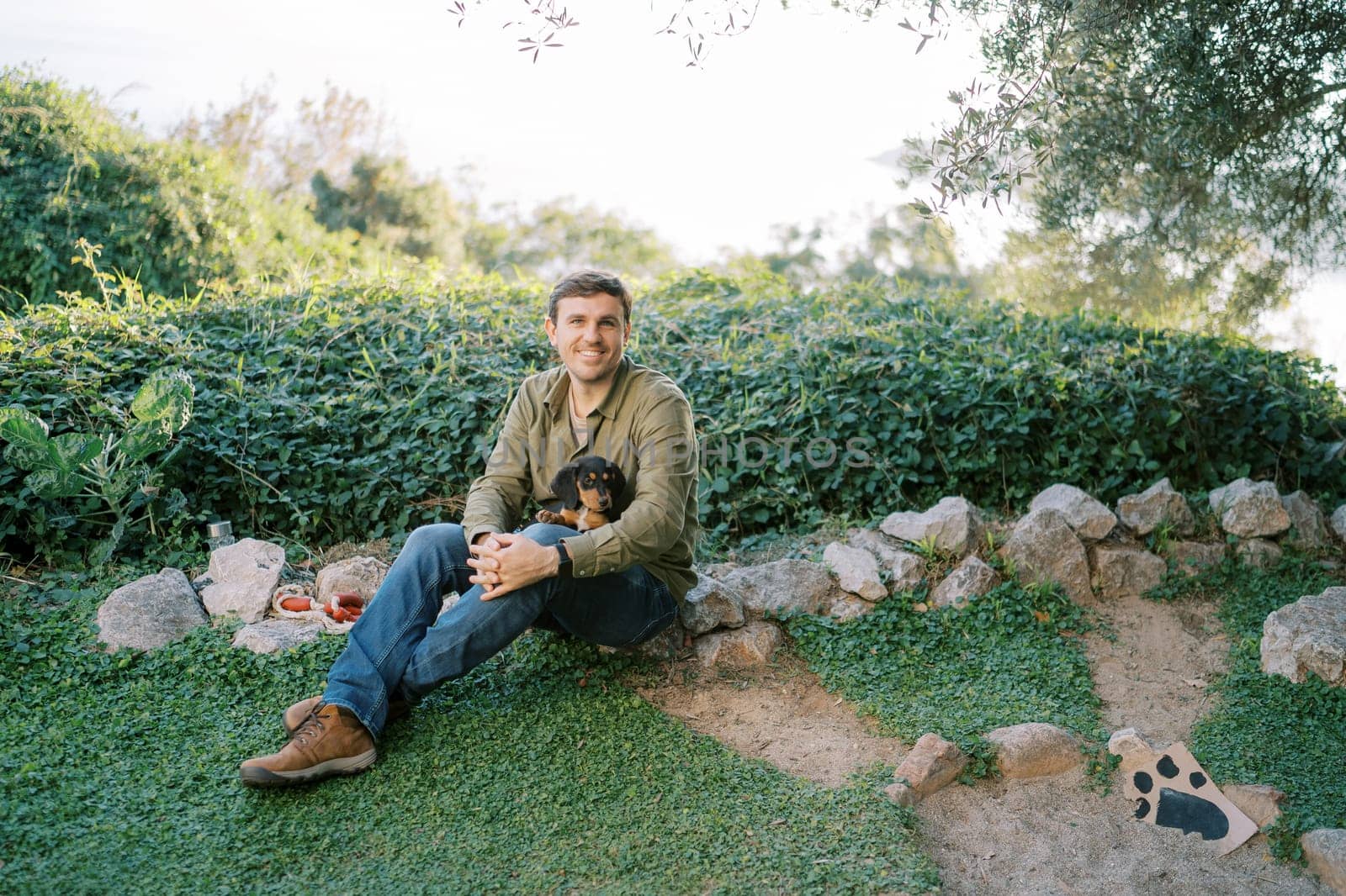 Smiling guy with a puppy on his knees sits on green grass near the bushes. High quality photo