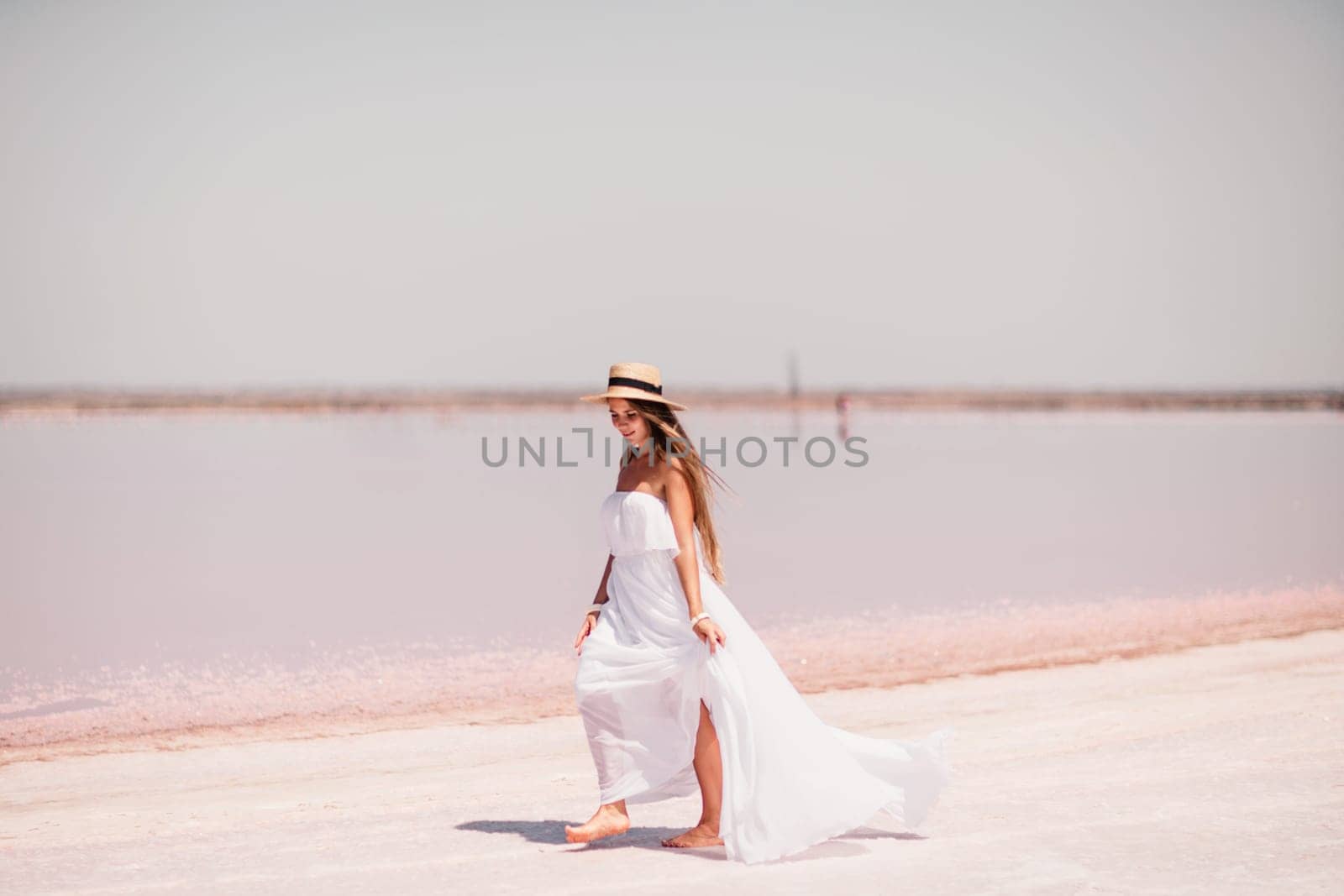 Woman in pink salt lake. She in a white dress and hat enjoys the scenic view of a pink salt lake as she walks along the white, salty shore, creating a lasting memory