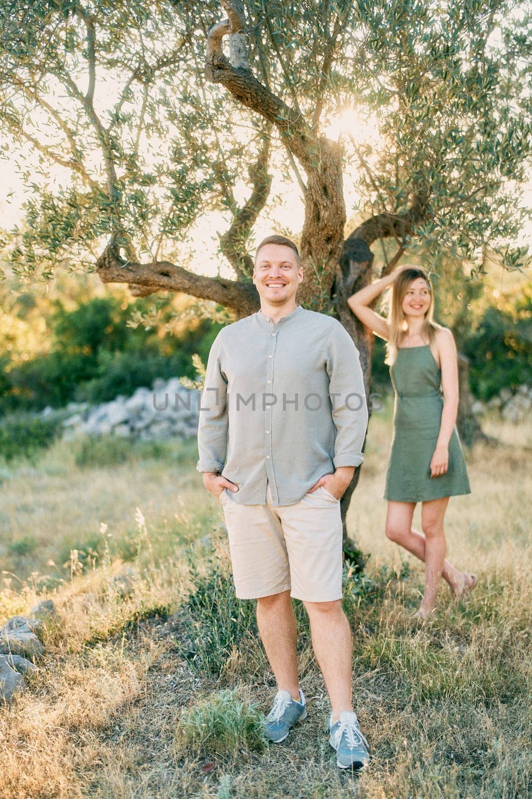 Girl stands leaning her elbow on a tree behind smiling guy with his hands in his pockets. High quality photo
