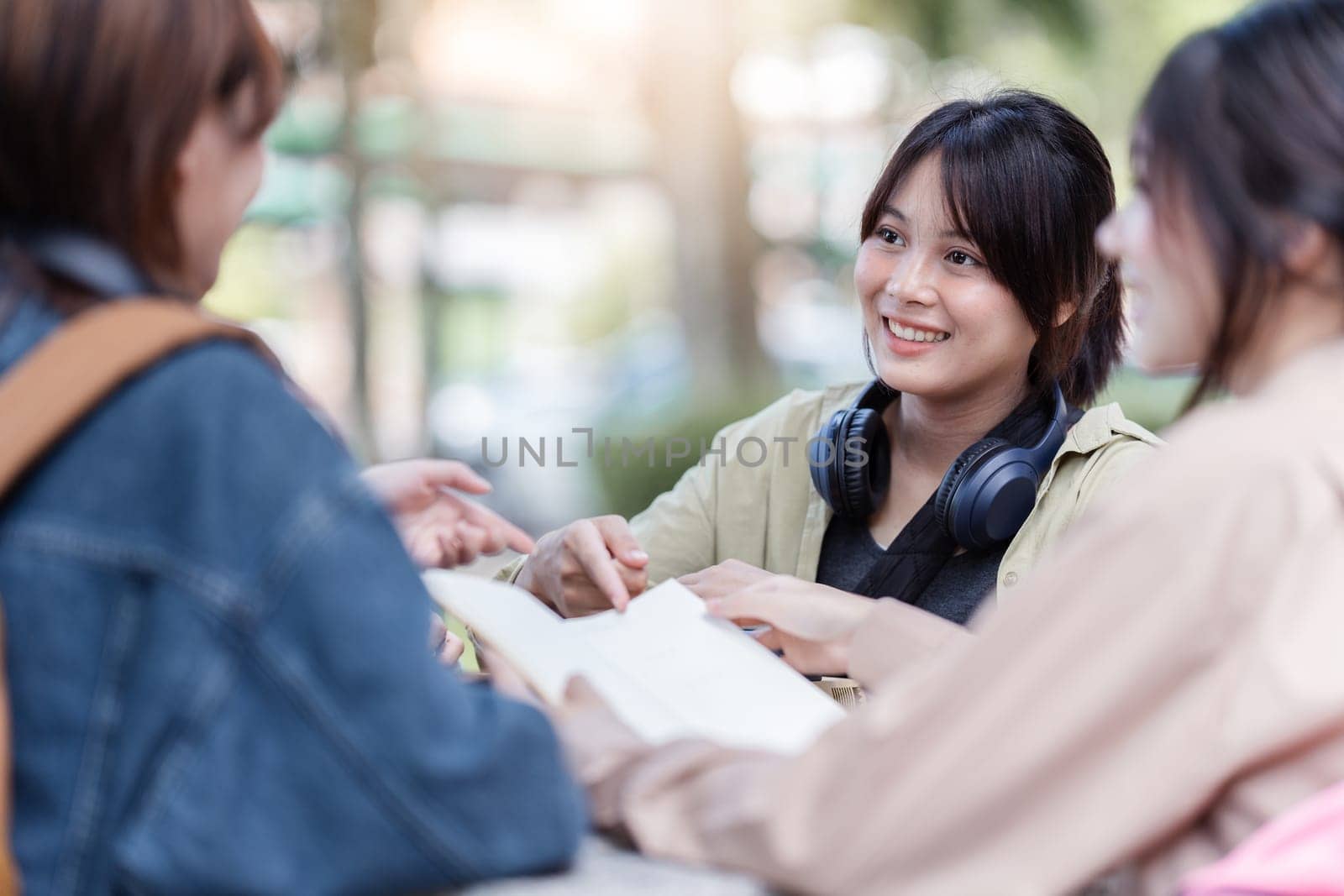 Group of young Asian college students sitting on a bench in a campus relaxation area, talking, sharing ideas, doing homework or tutoring for the exam together.