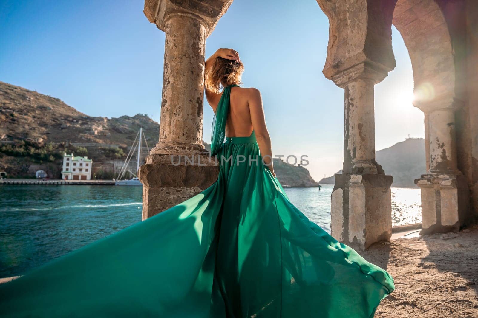 Woman dress sea columns. Rear view of a happy blonde woman in a long mint dress posing against the backdrop of the sea in an old building with columns