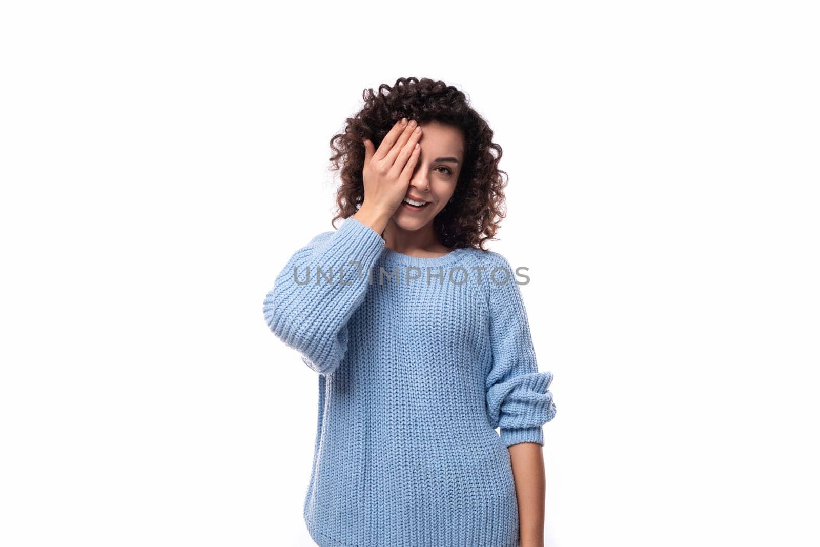 positive young lady with black curly hair dressed in a light blue sweater posing on a white background.