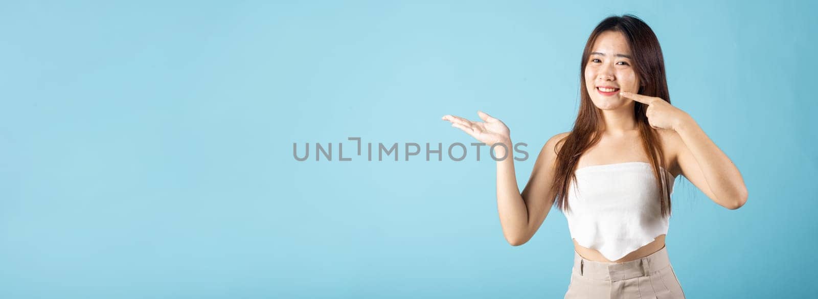 Asian young woman pointing to strong white teeth and open hand palm for display cosmetics product, female point fingers to smiling mouth studio shot isolated on blue background, Dental health concept