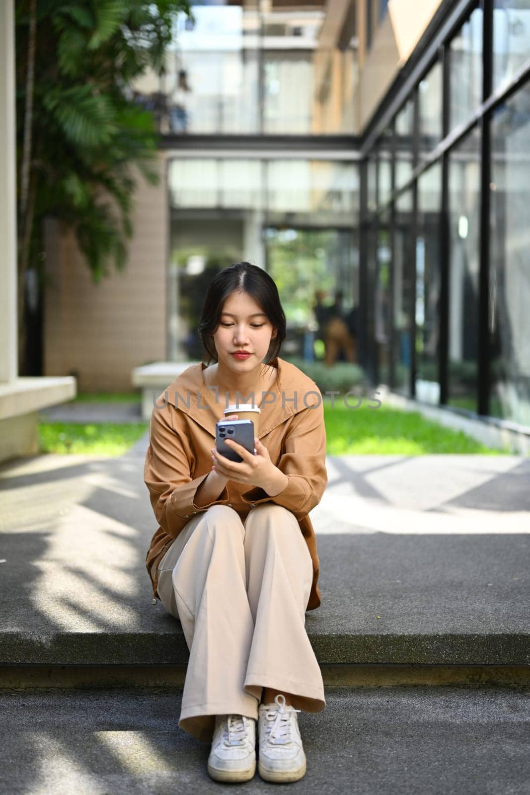 Portrait of pretty young Asian woman sitting front of business center and using mobile phone by prathanchorruangsak