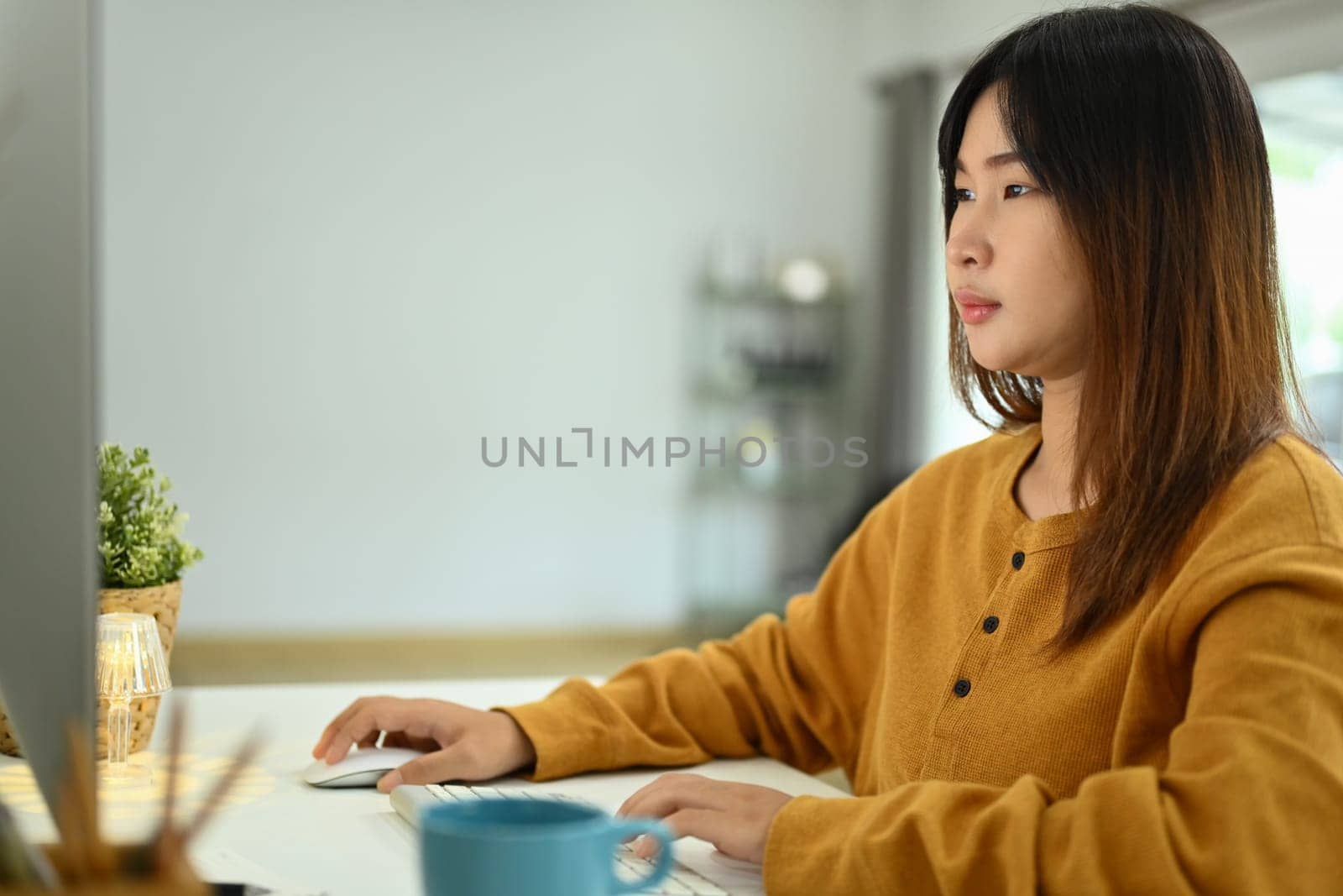 Concentrate young Asian female looking at computer screen working online at home by prathanchorruangsak