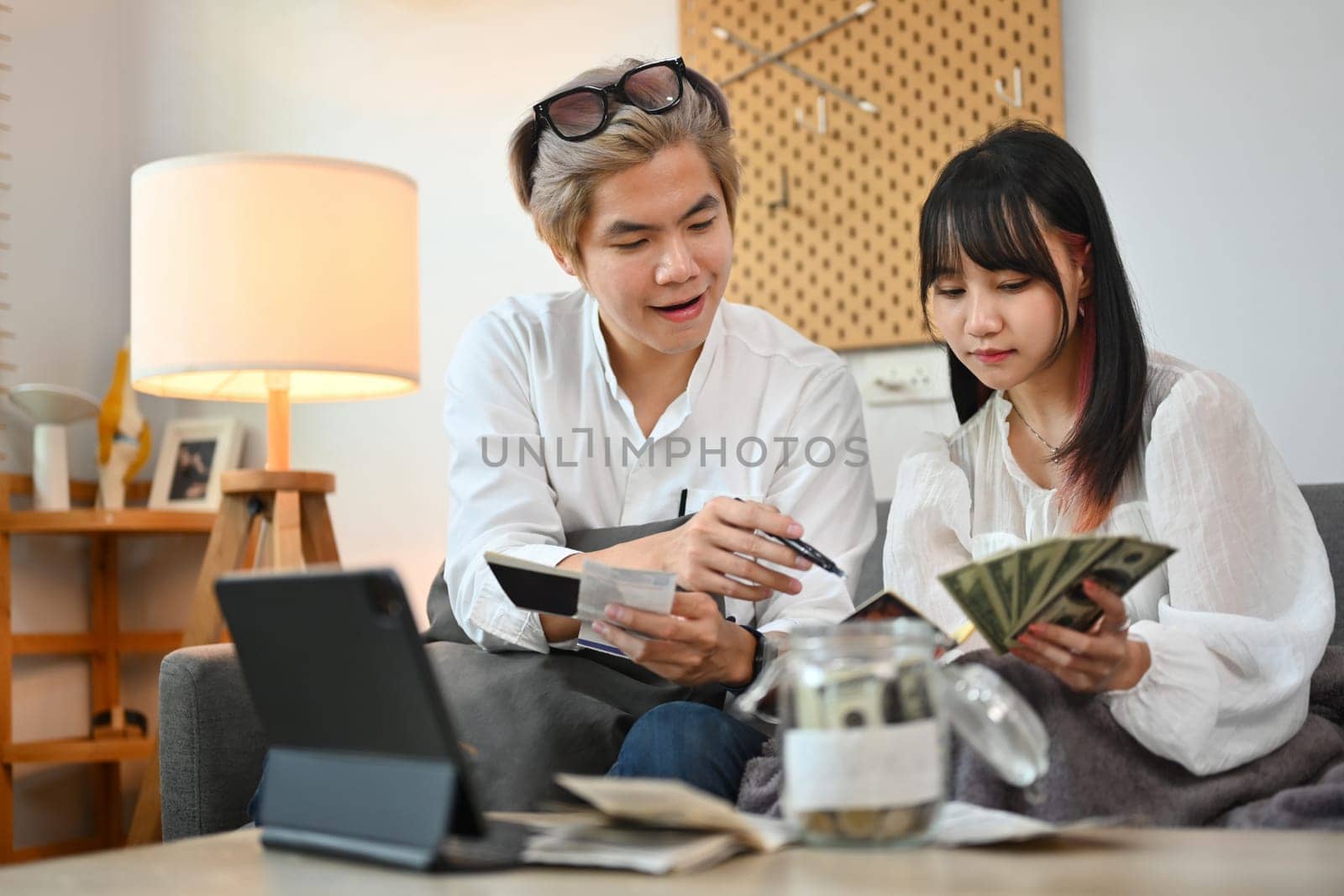 Smiling young couple sitting in living room calculating expenses, managing finances and save money for future.