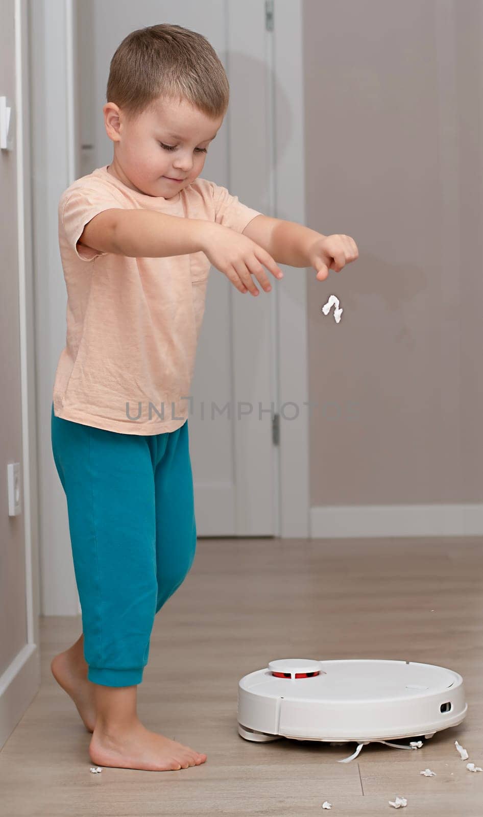 Cleaning concept. A small funny Caucasian boy, 4 years old, throws torn paper on the floor for cleaning by a white robot vacuum cleaner. Plays happily in the home interior. by ketlit
