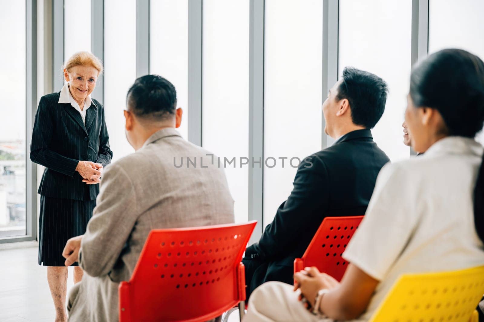 In a dynamic office meeting, a confident senior corporate coach delivers a seminar to a business team. The female director, CEO, and employees actively participate, discussing projects and guiding. by Sorapop