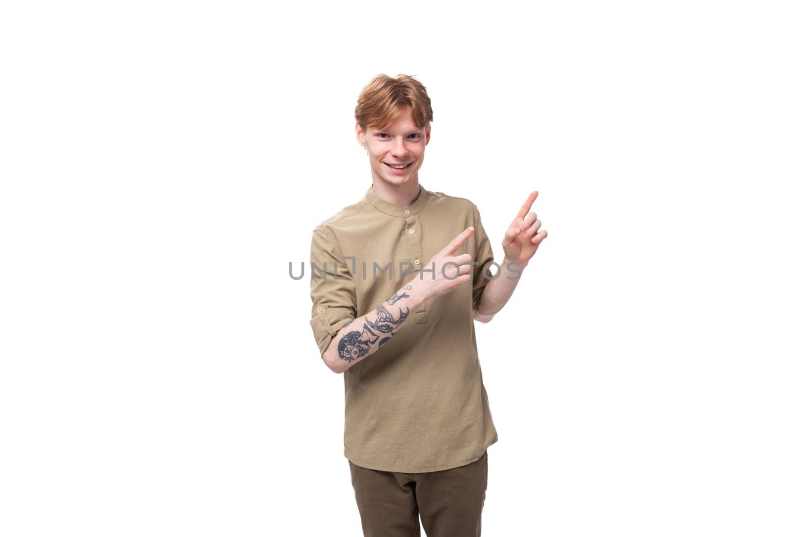 young surprised red-haired caucasian guy dressed in a khaki short sleeve shirt points to the side.