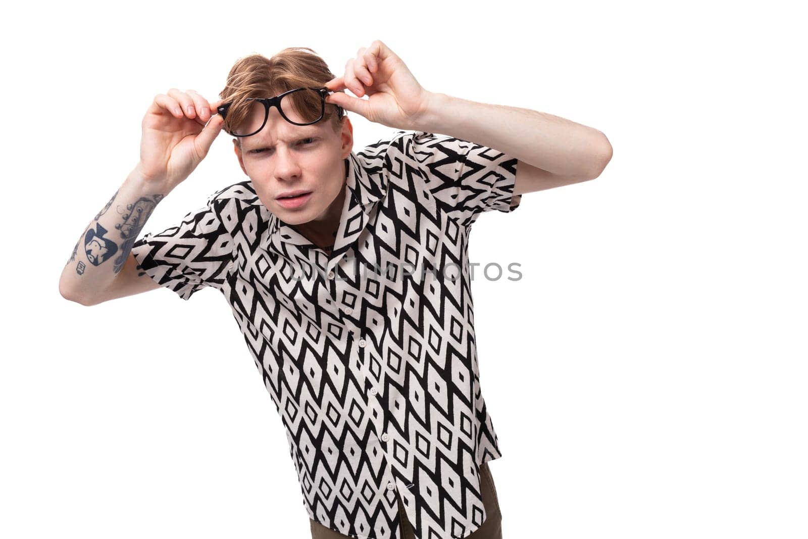 European young man with golden hair and a tattoo on his arms dressed in a summer black and white shirt is experiencing discomfort from wearing glasses.