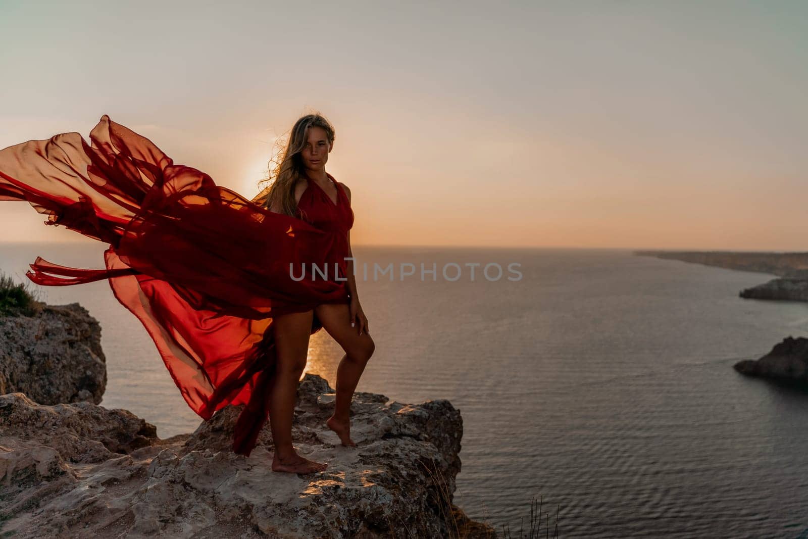 Woman sunset sea red dress, side view a happy beautiful sensual woman in a red long dress posing on a rock high above the sea on sunset