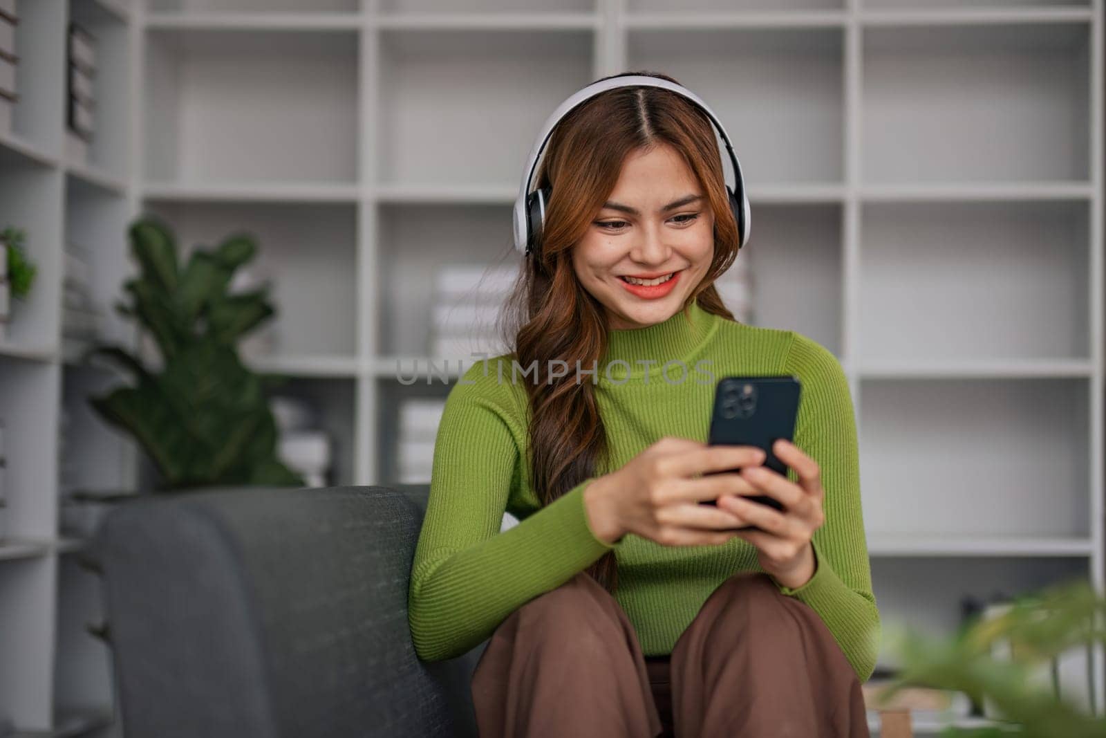 Happy Asian woman listening to music on mobile phone while sitting on sofa at home Relaxed smiling girl with headphones in the morning It's time to rest. by wichayada