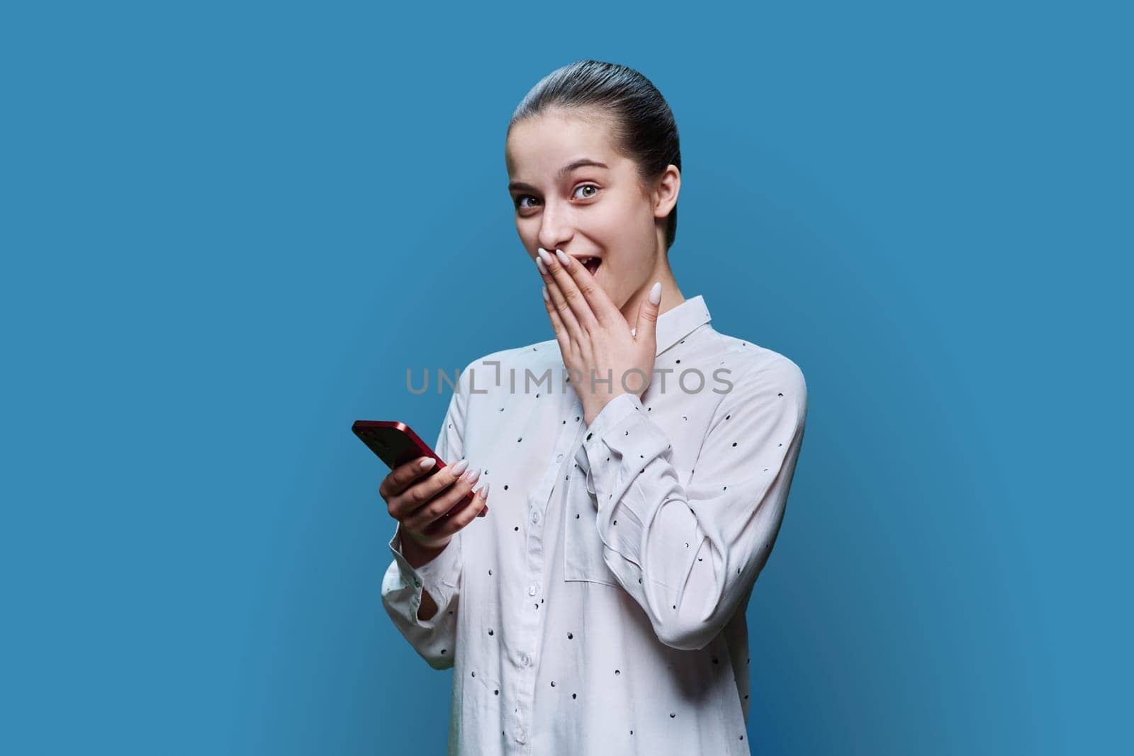 Surprised teenage student, girl 15, 16 years old using smartphone on blue studio background. Adolescence, internet technologies, mobile online applications apps services for learning leisure