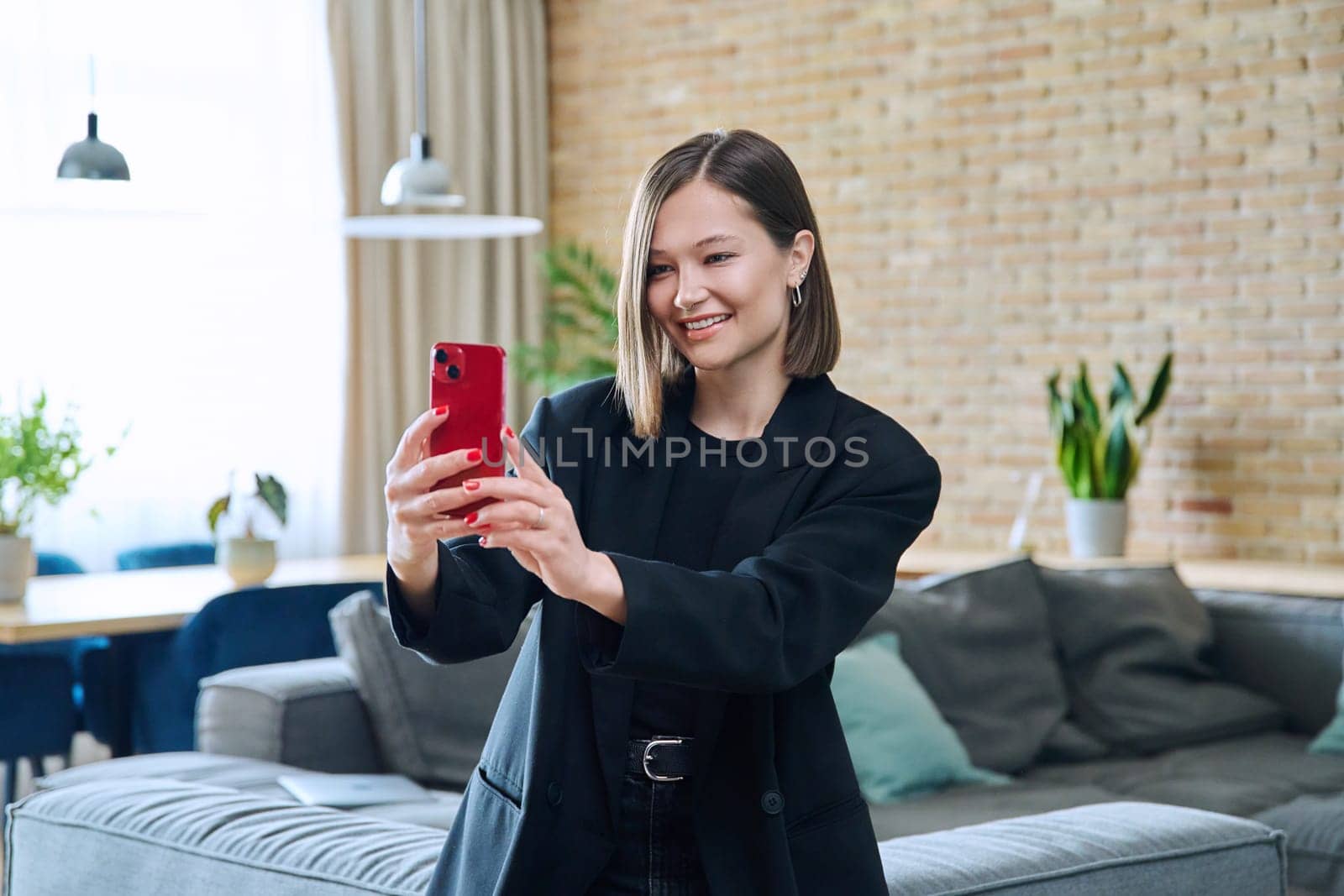 Young smiling joyful happy woman sitting on the sofa in the living room having a video call chat conference using a smartphone. Communication, leisure, home, lifestyle, youth, technology concept