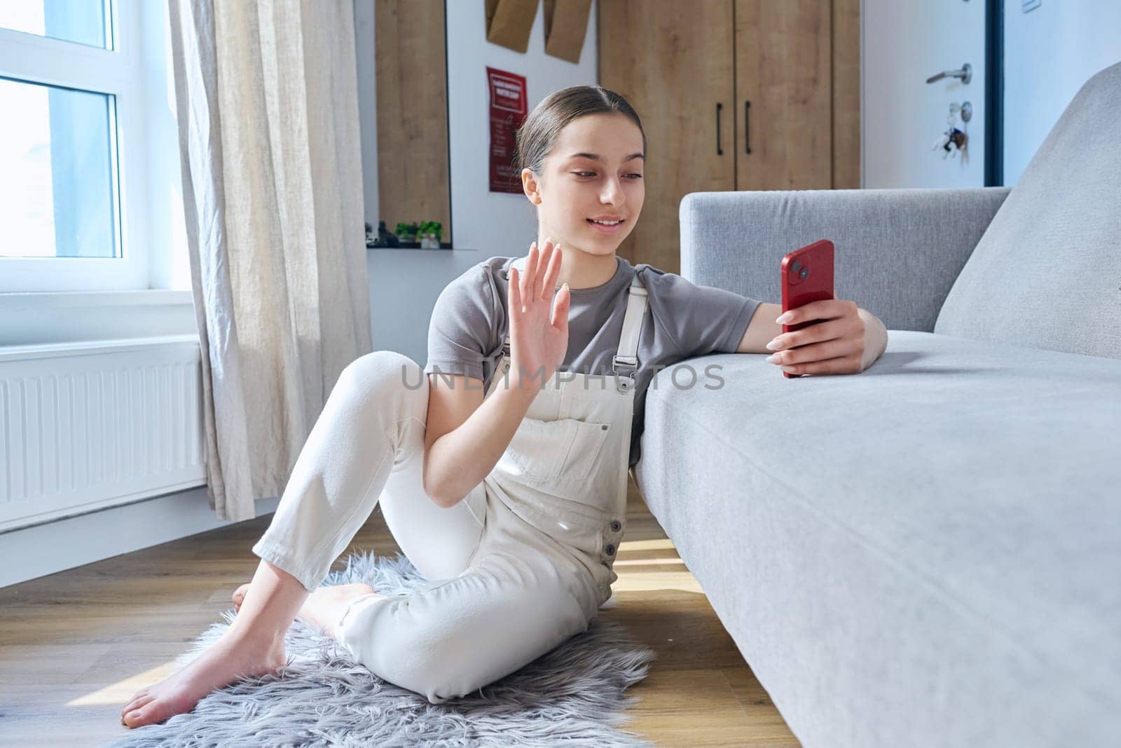 Teenager girl sitting at home on floor near sofa using smartphone for video call conference, waving hand at phone screen. Technology for learning leisure communication, modern lifestyle, adolescence