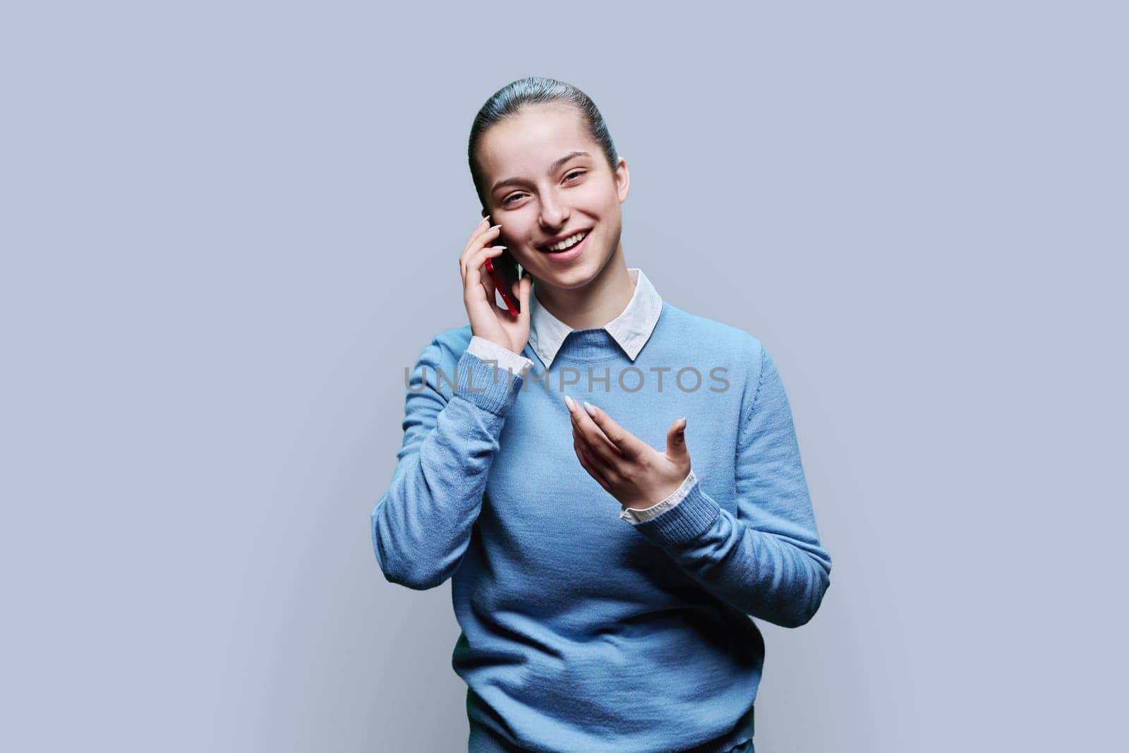 Joyful emotional surprised happy cheerful teenage student, girl 15, 16 years old talking on mobile phone on gray studio background. Lifestyle, communication, adolescence concept