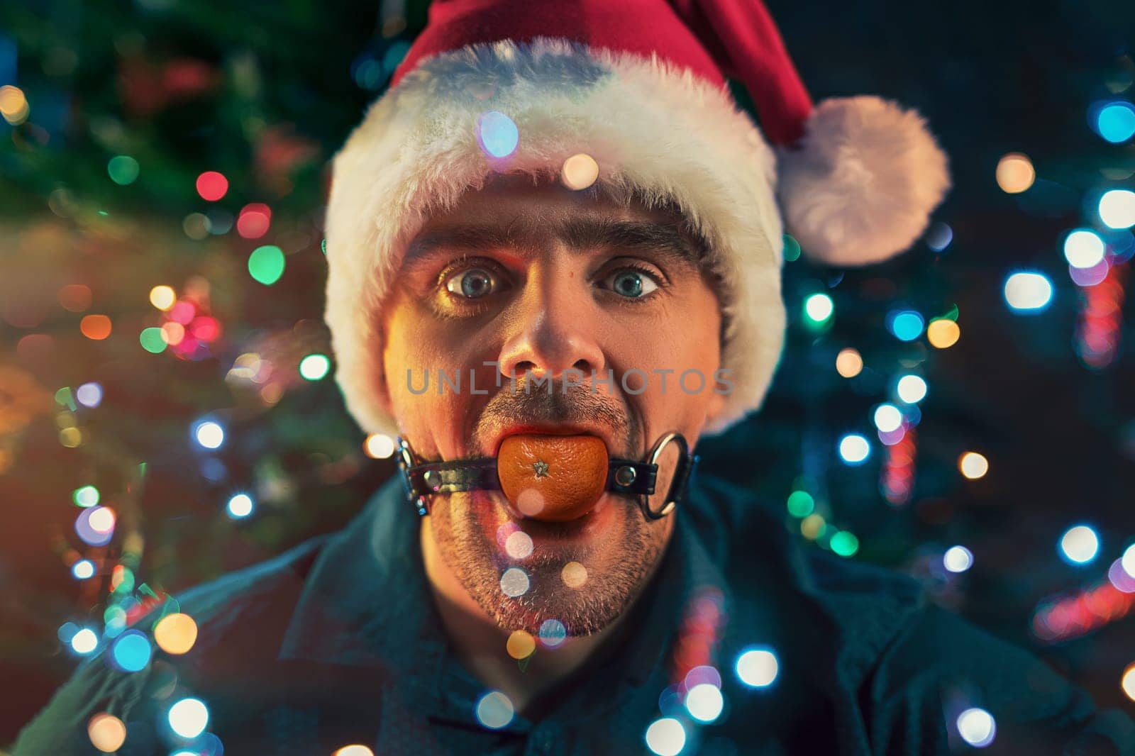Man with mandarin in his mouth as BDSM sex toys on the Christmas tree background. Concept of fatigue of celebrating New Year's Eve.