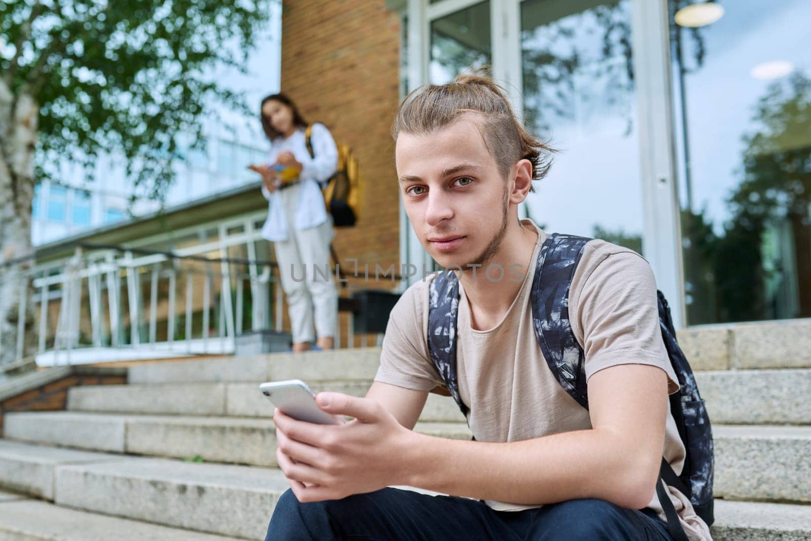 Hipster guy smiling teenager student 18, 19 years old with backpack smartphone looking at camera, sitting on steps of outdoor educational building. Youth, education, lifestyle, technology