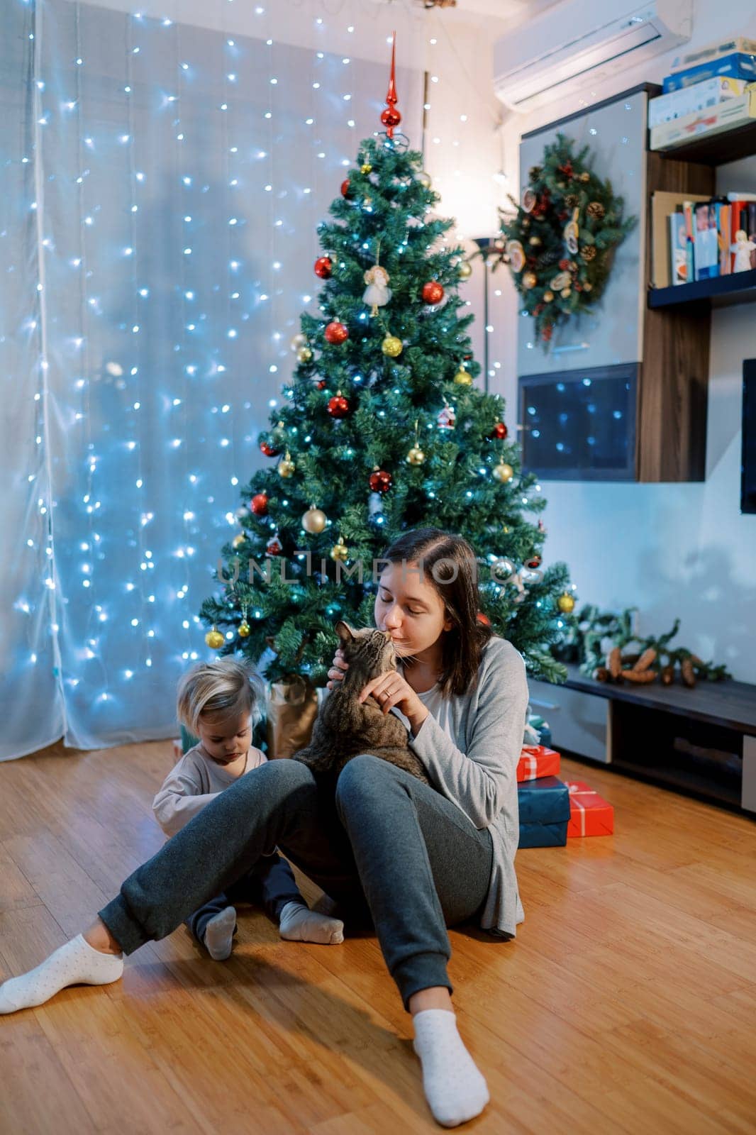Mom pets a cat sitting on the floor with a little girl near the Christmas tree. High quality photo