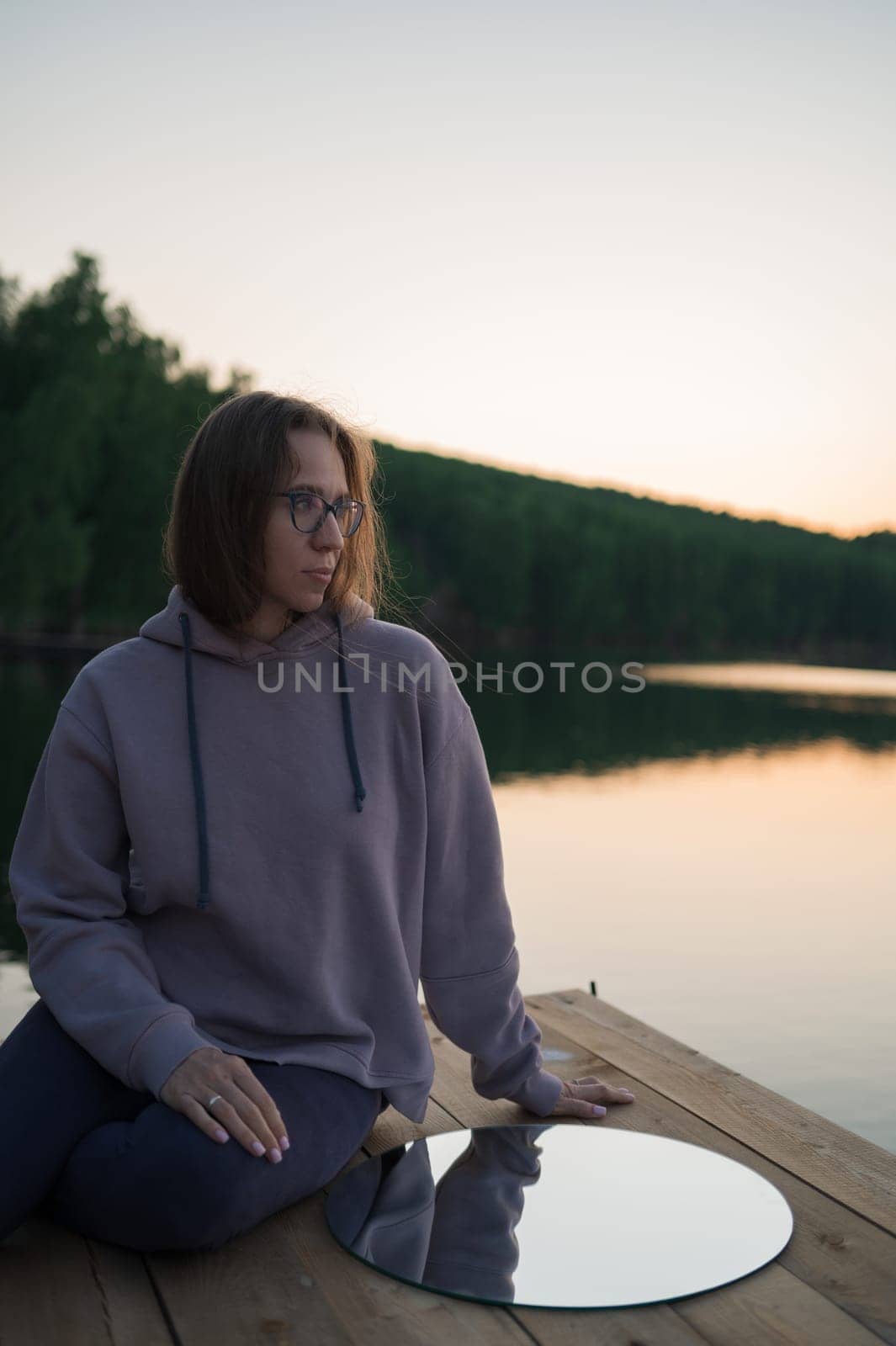 Woman sitting on the pier at lake with circle mirror, summer sunset