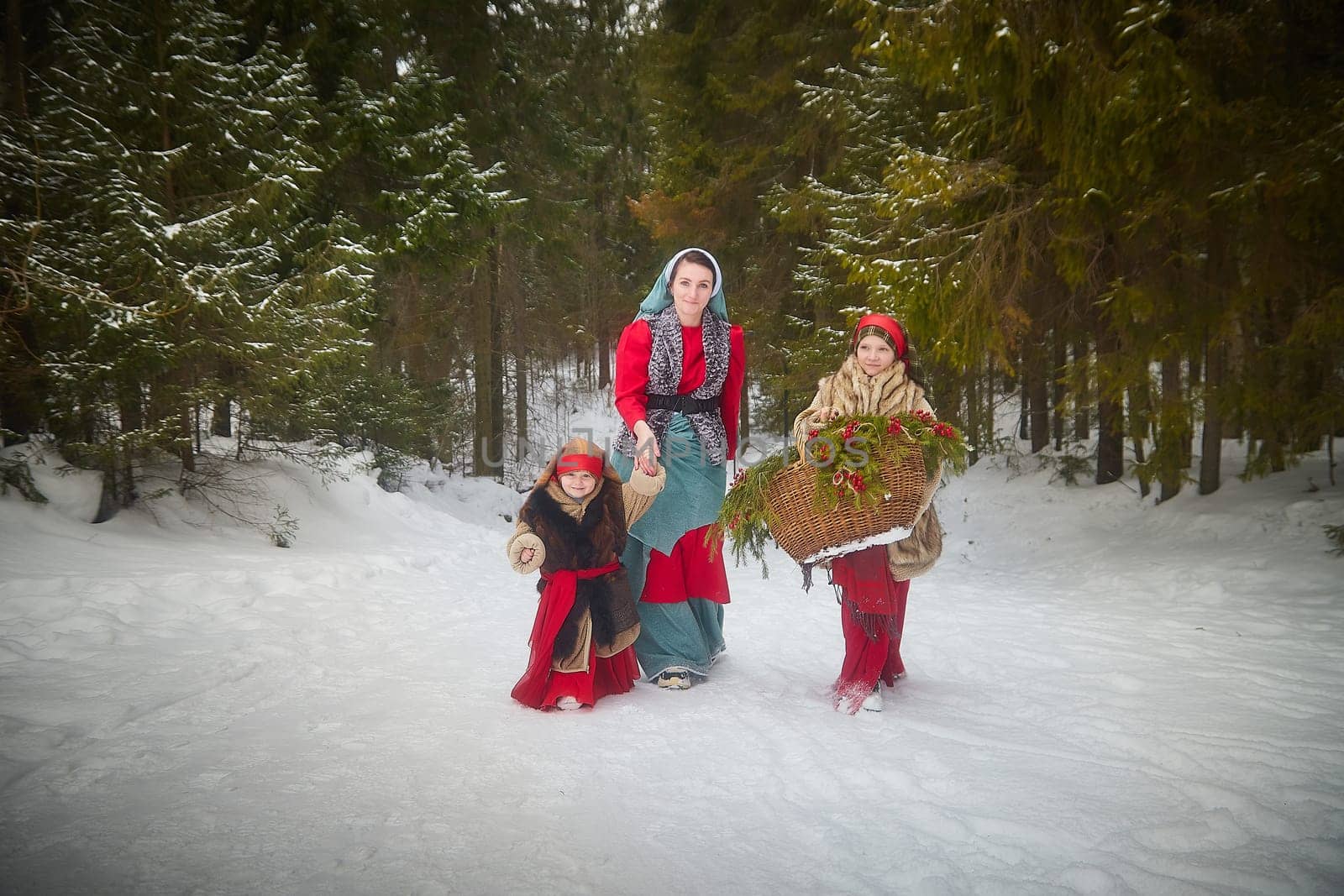 Family with mother, teenage girl, and little daughter dressed in stylized medieval peasant clothing in winter forest. Woman and her daughters pose for fairytale photoshoot in nature on cold day by keleny