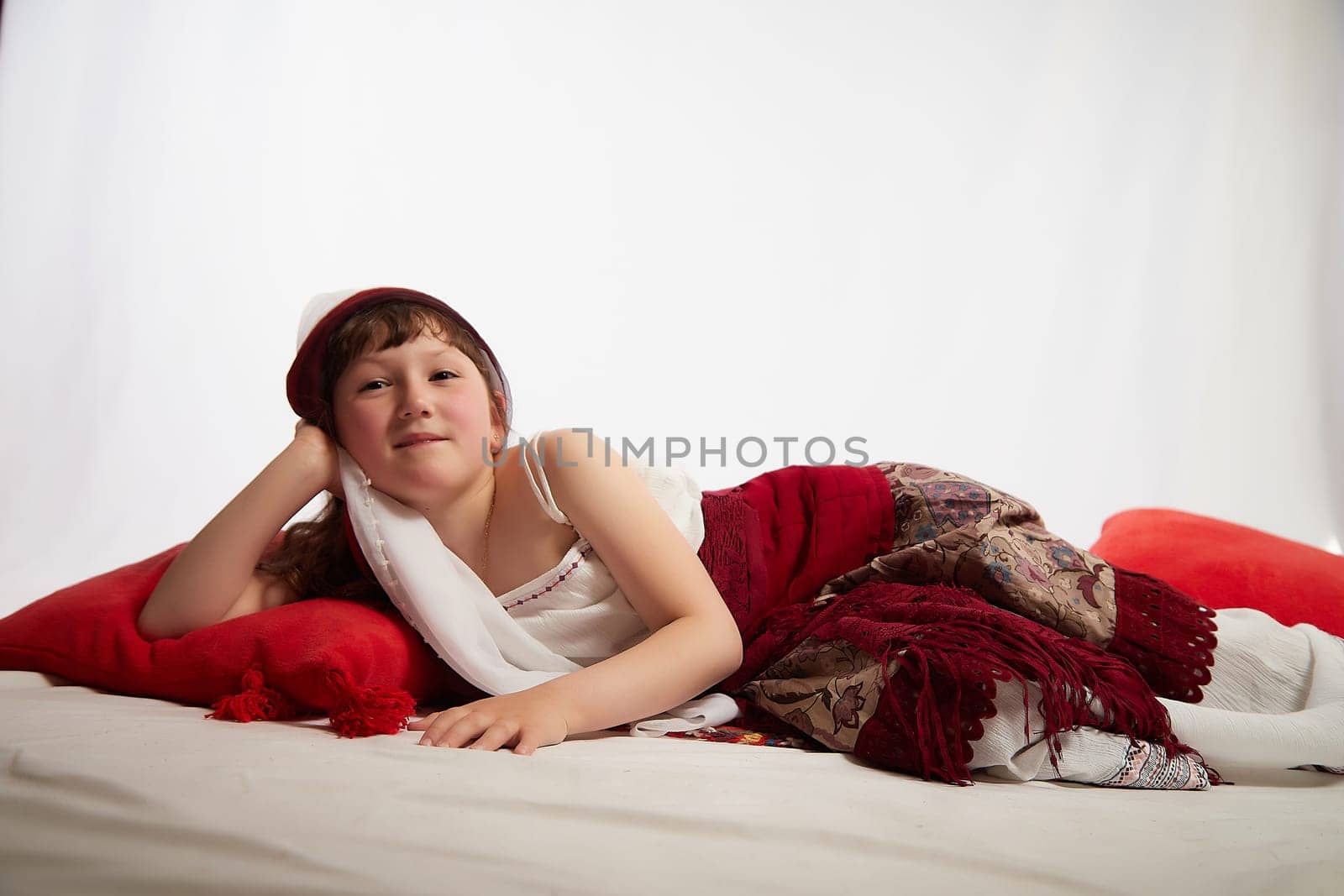 Portrait of Little girl in a stylized Tatar national costume having rest with a red pillow on a white background in the studio. Photo shoot of funny young teenager who is not a professional model