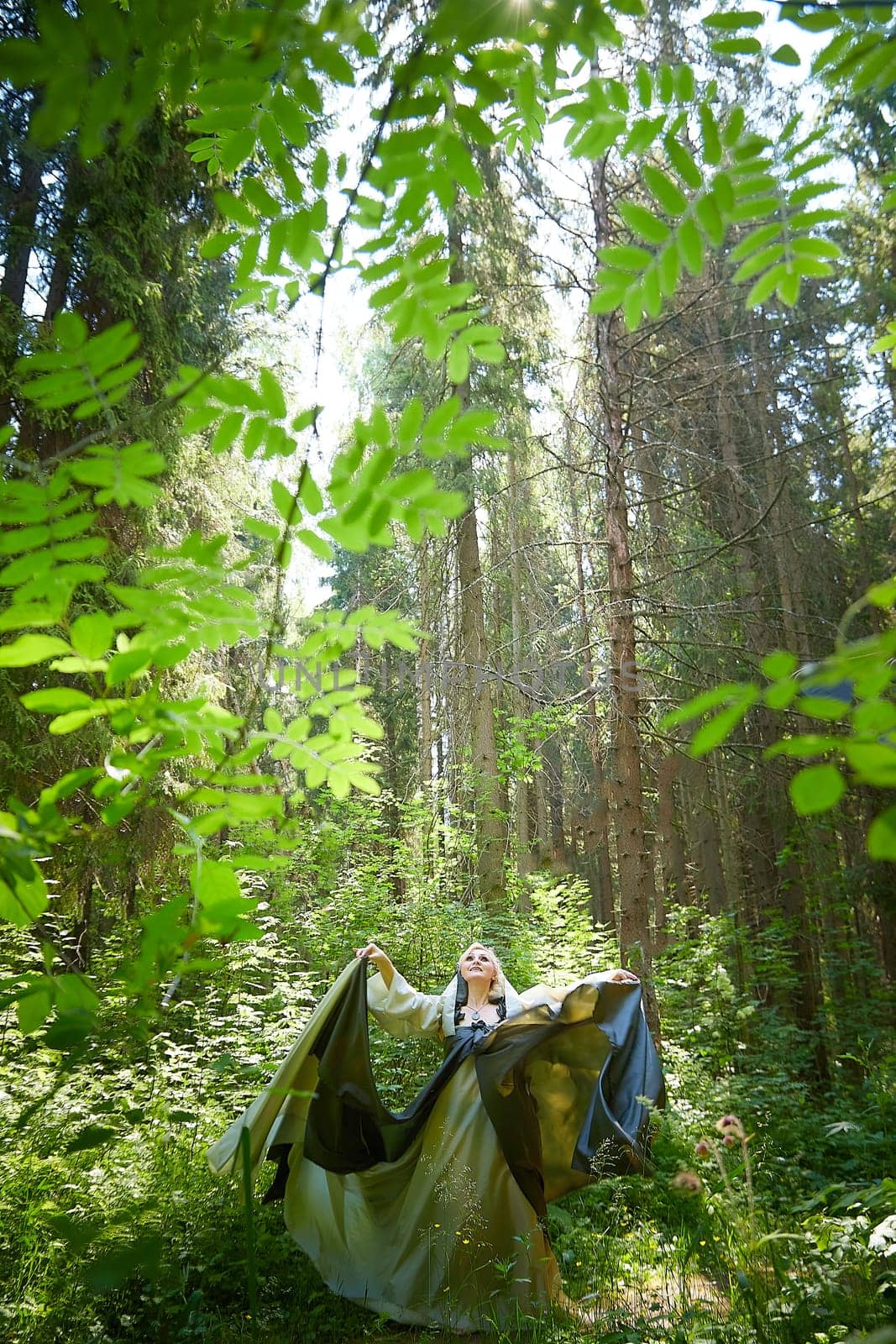 Adult mature woman 40-60 in a green long fairy dress in forest. Photo shoot in style of dryad and queen of nature. Fairy in beautiful green summer forest. Concept of caring for nature