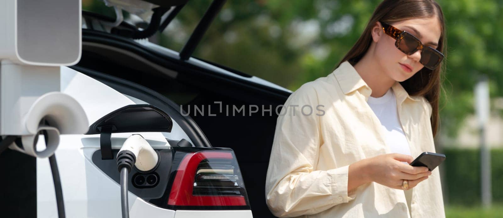 Young woman use smartphone to pay for electricity at public EV car charging station green city park. Modern environmental and sustainable urban lifestyle with EV vehicle. Panorama Expedient