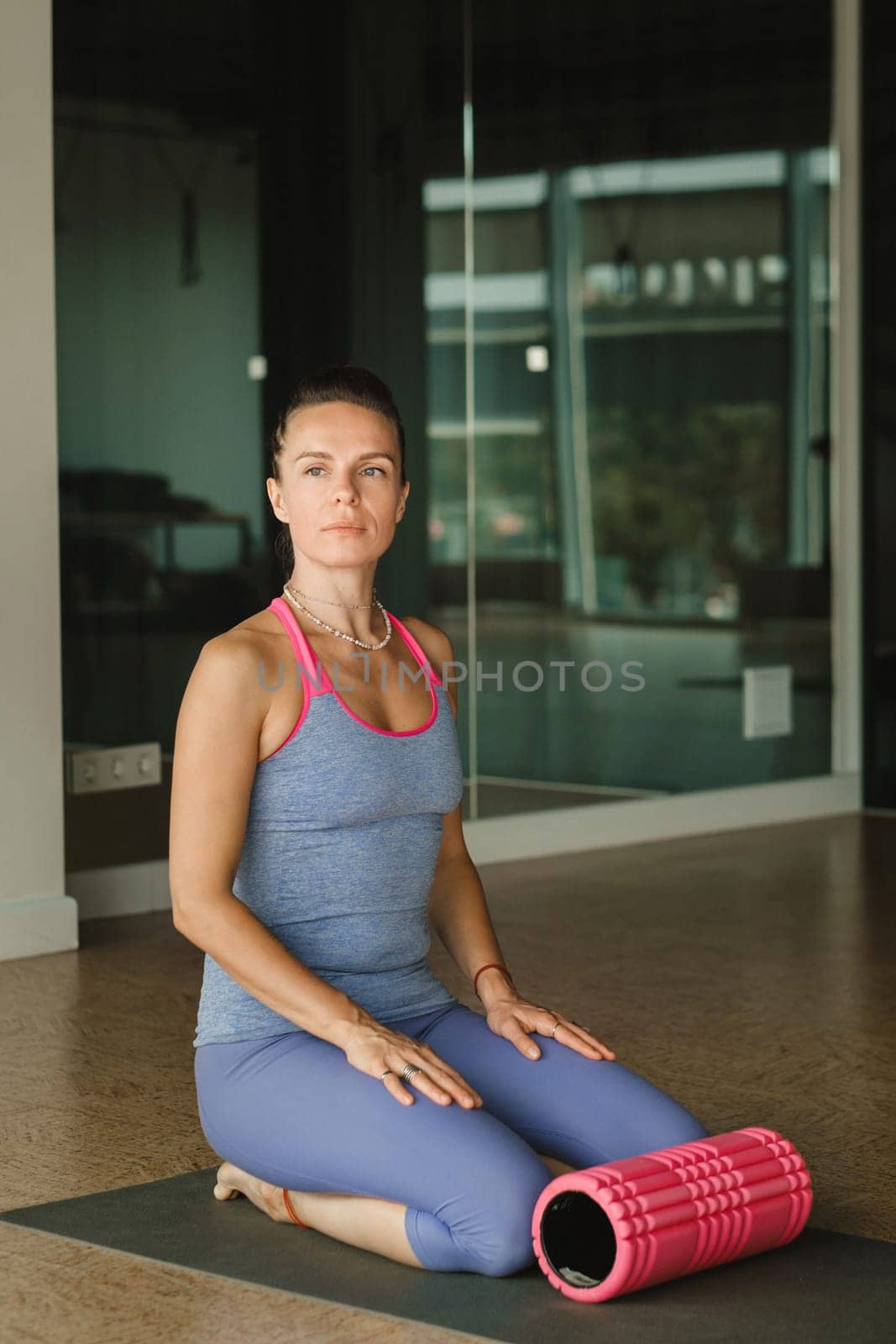 A slender girl is sitting on a yoga mat with a big massage roller in the fitness room by Lobachad