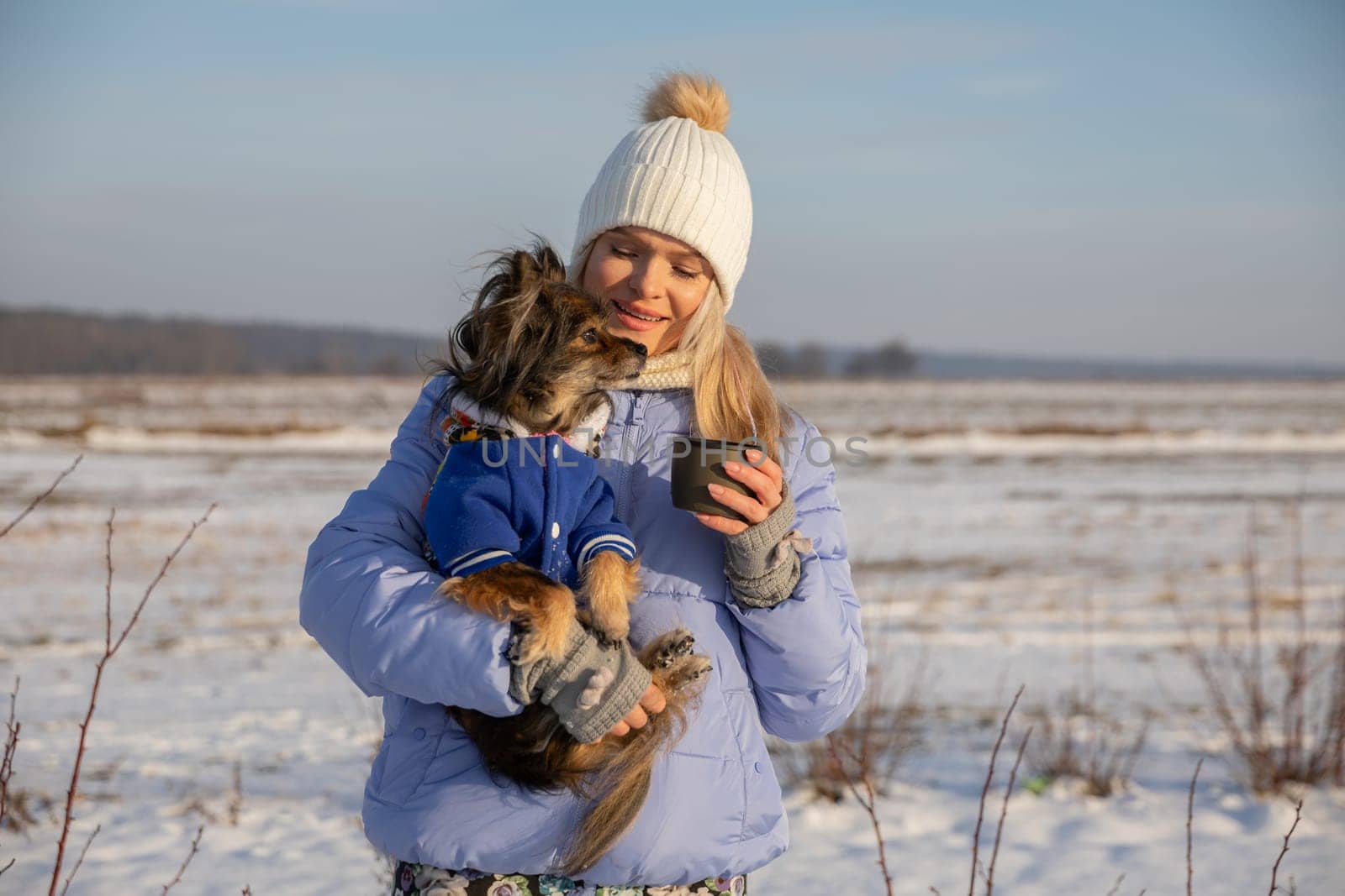 Blurred background. In the background you can see a snowy field and leafless bushes. In the foreground a young girl is holding a small breed dog in her arms. The dog is wearing a jacket.