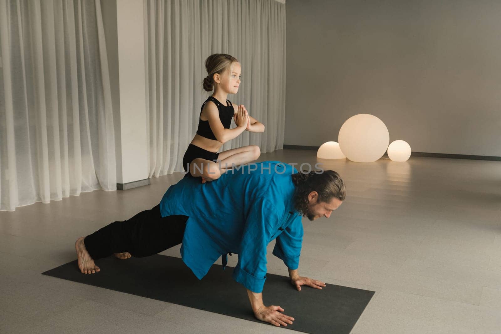 a child in the lotus position sits on the back of a yoga coach during training by Lobachad