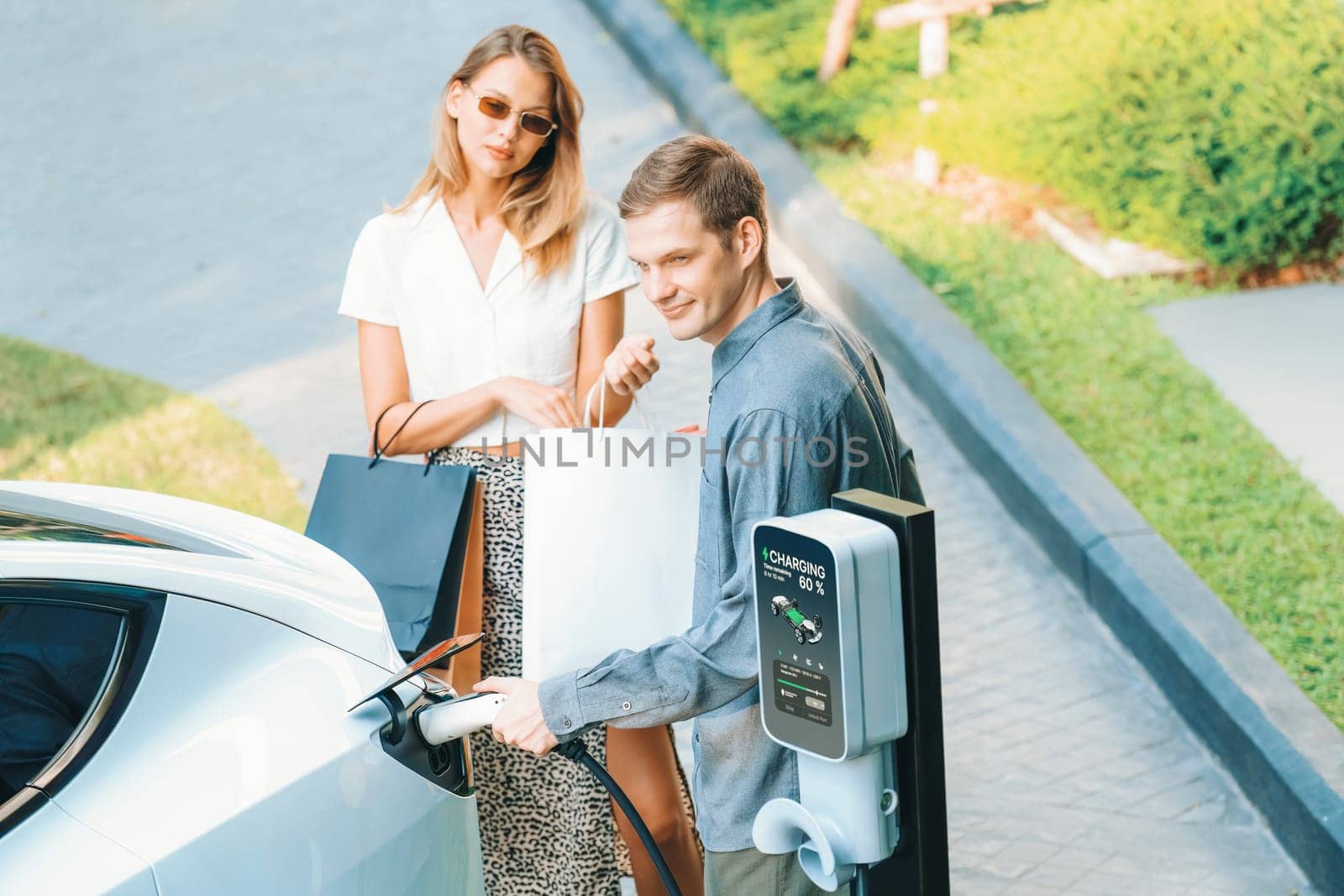 Young couple travel with EV electric car charging in green sustainable city outdoor garden in summer shows urban sustainability lifestyle by green clean rechargeable energy of electric vehicle innards