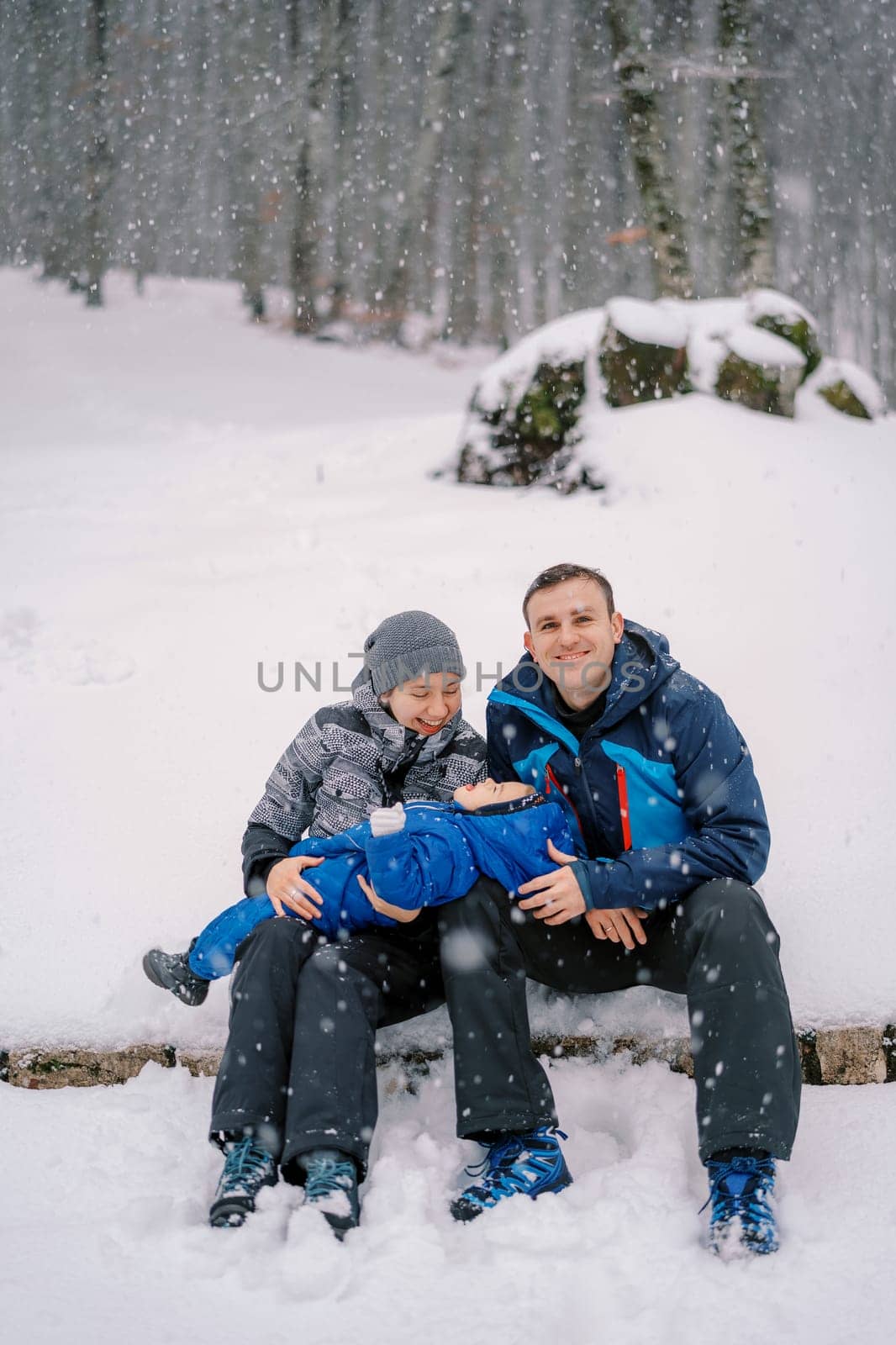 Little girl lies on the knees of smiling mom and dad and catches snowflakes in her mouth in the forest. High quality photo