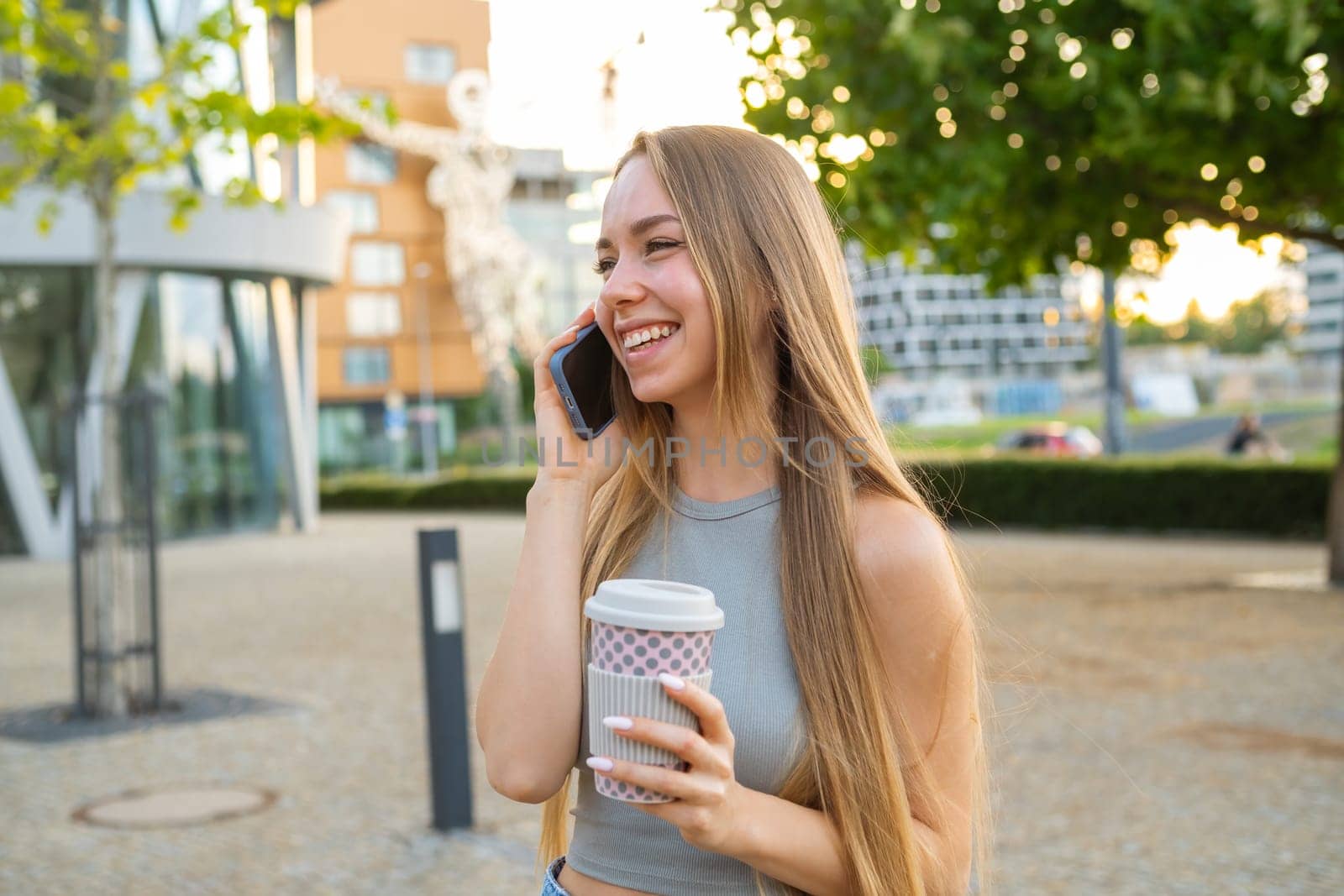 Cheerful young woman with coffee cup in hand laughs talking on phone with boyfriend. Lady spends time drinking coffee in city park after work