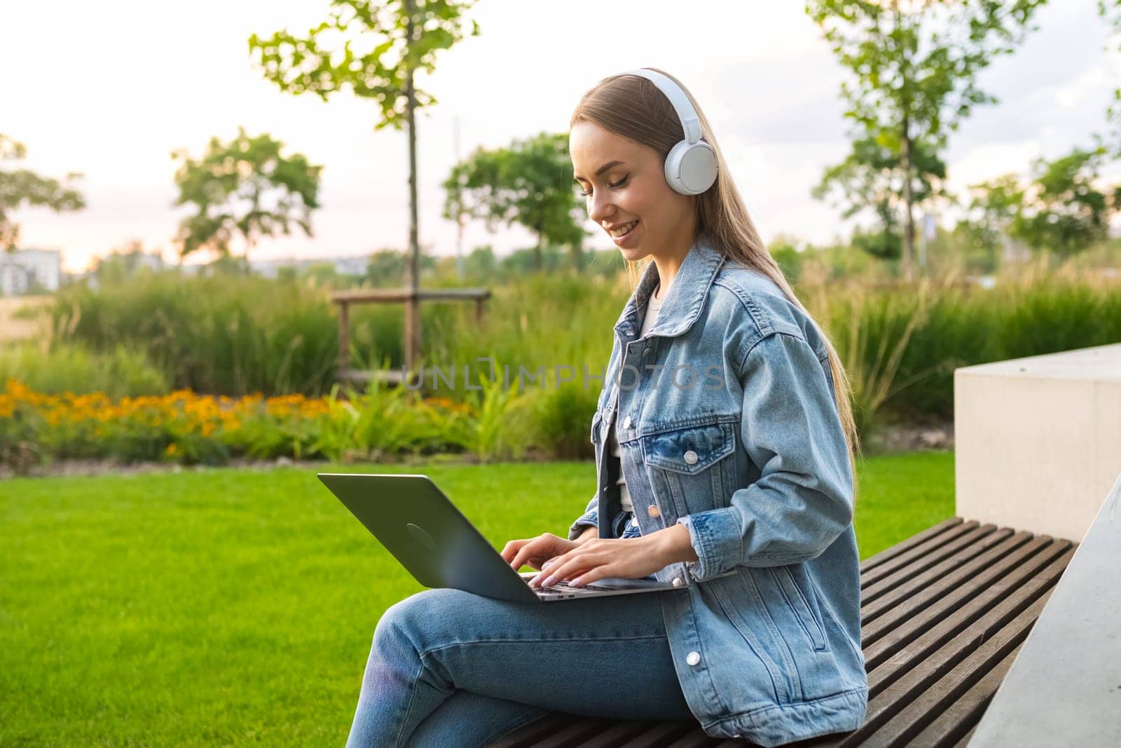 Young blonde woman with laptop on lap looks in camera sitting on park bench in evening. Lady in denim jacket rests in park after work day