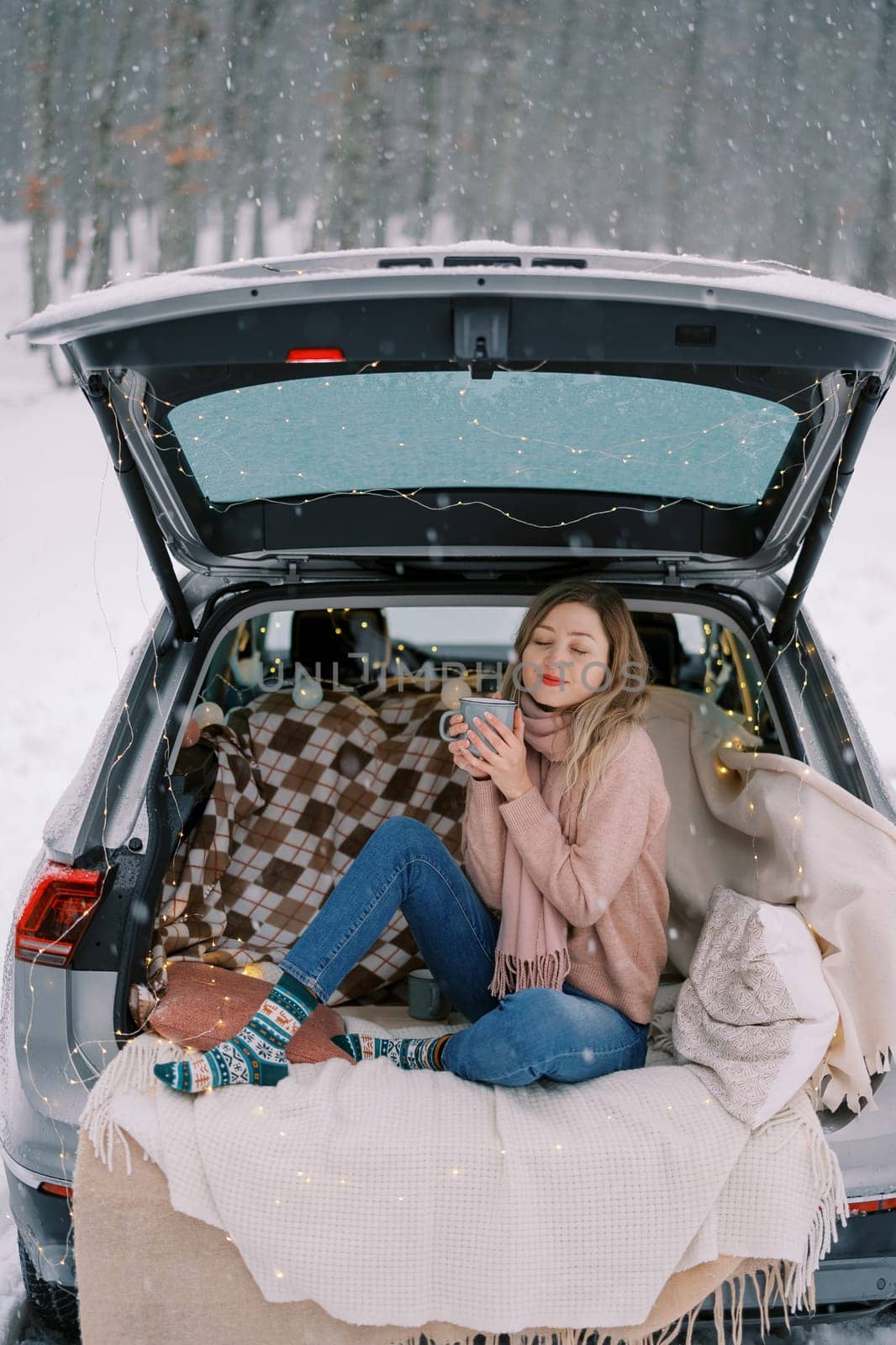 Young woman sits with a mug of coffee squinting in pleasure in the trunk of a car in a winter forest by Nadtochiy